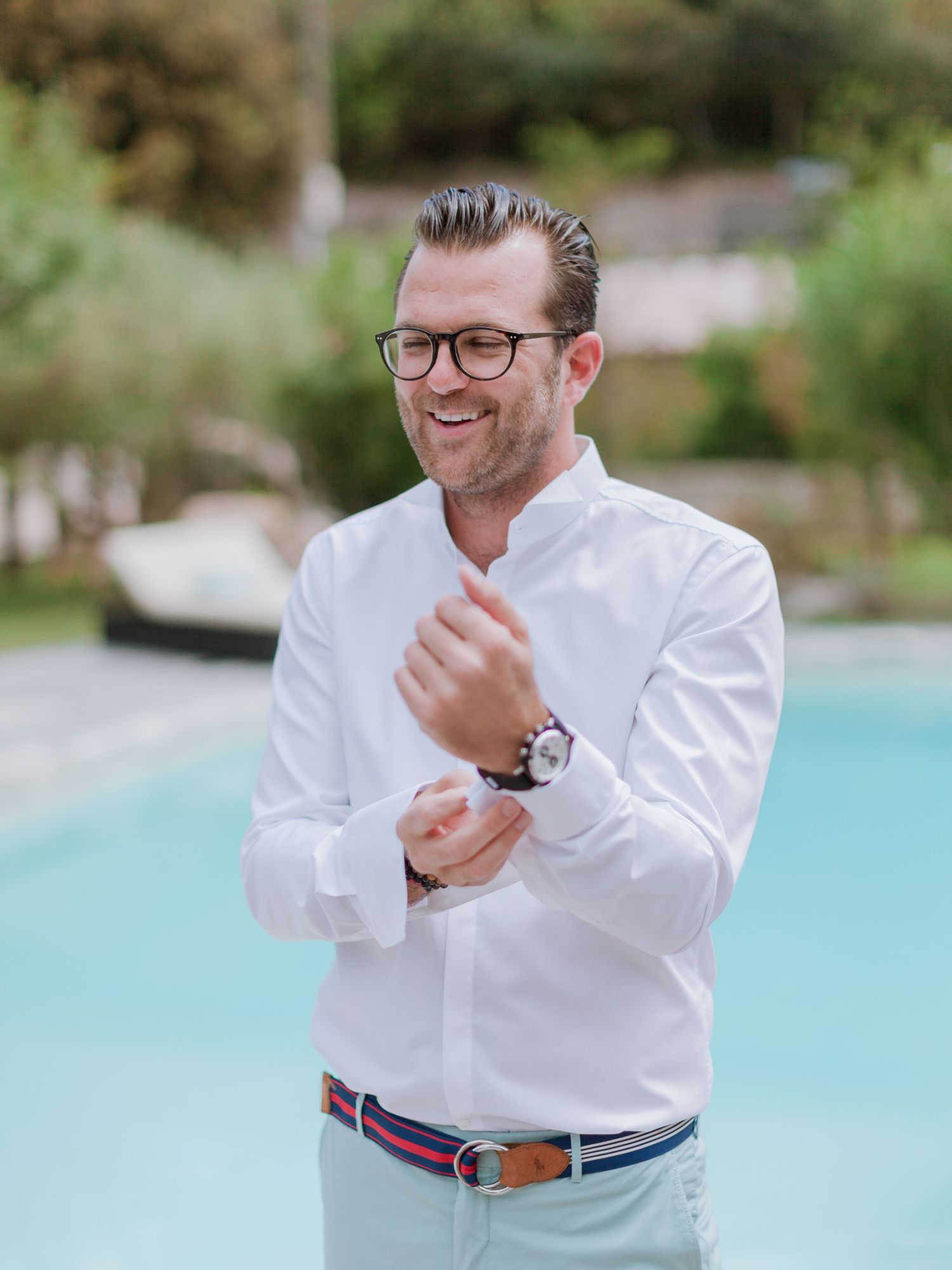 Un homme souriant, vêtu d'une chemise blanche et portant des lunettes, ajuste ses boutons de manchette au bord d'une piscine