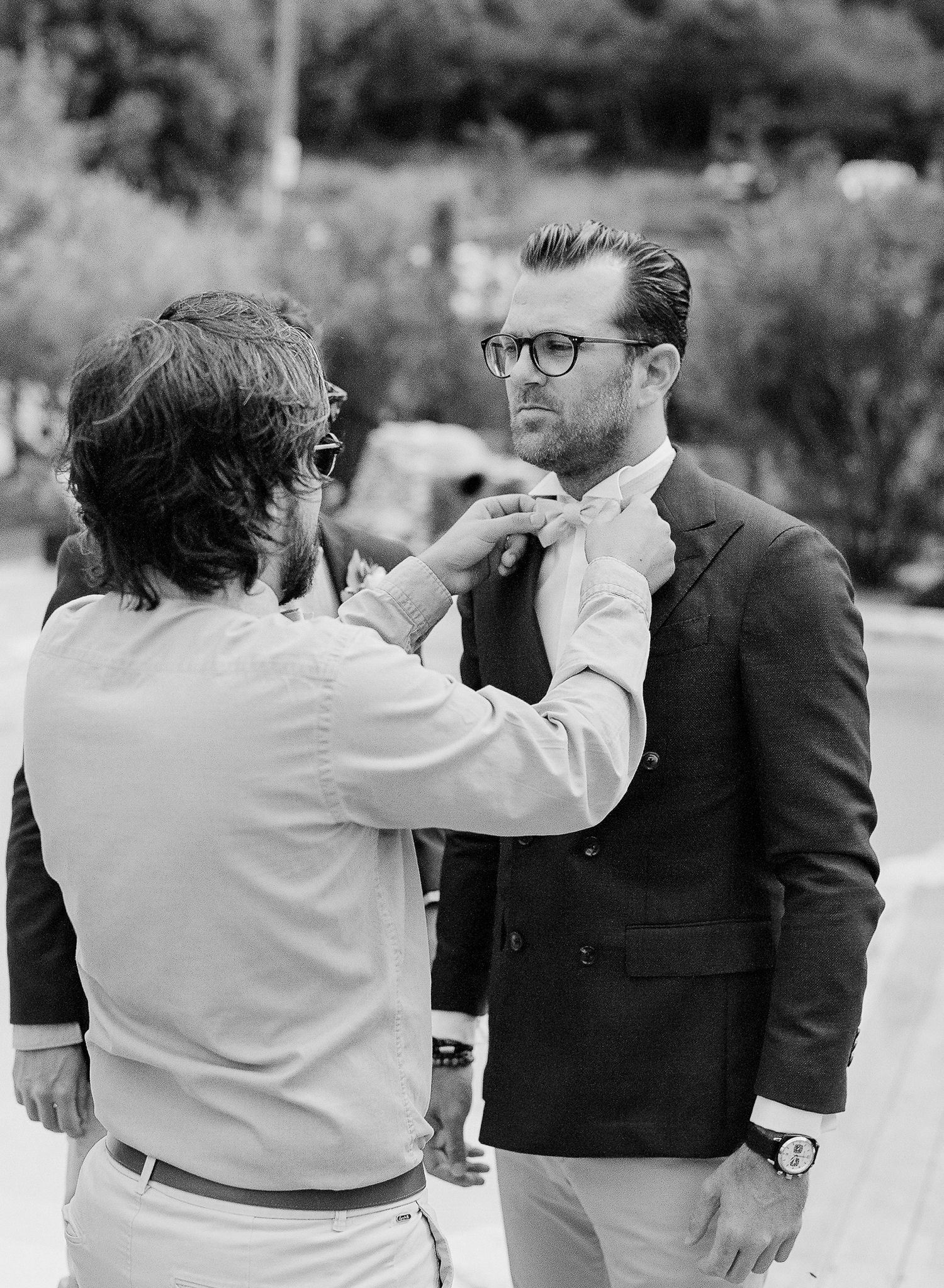 Photo en noir et blanc d'un homme portant des lunettes et un blazer foncé, se faisant ajuster son nœud papillon à l'extérieur.