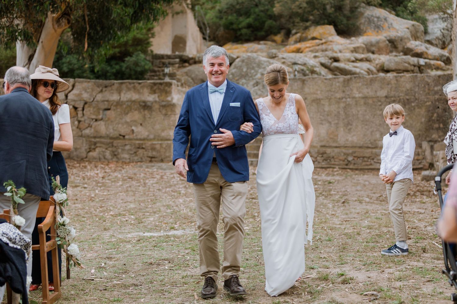 Les mariés descendent l'allée de mariage en plein air dans un lieu champêtre, l'homme en blazer bleu, la femme en robe de dentelle blanche.