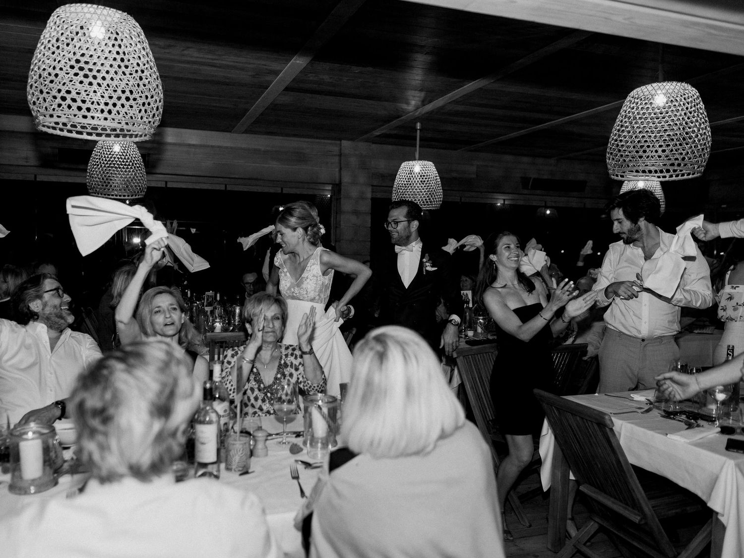 Photo en noir et blanc d'une réception de mariage montrant un couple debout parmi les invités en fête, sous des guirlandes lumineuses