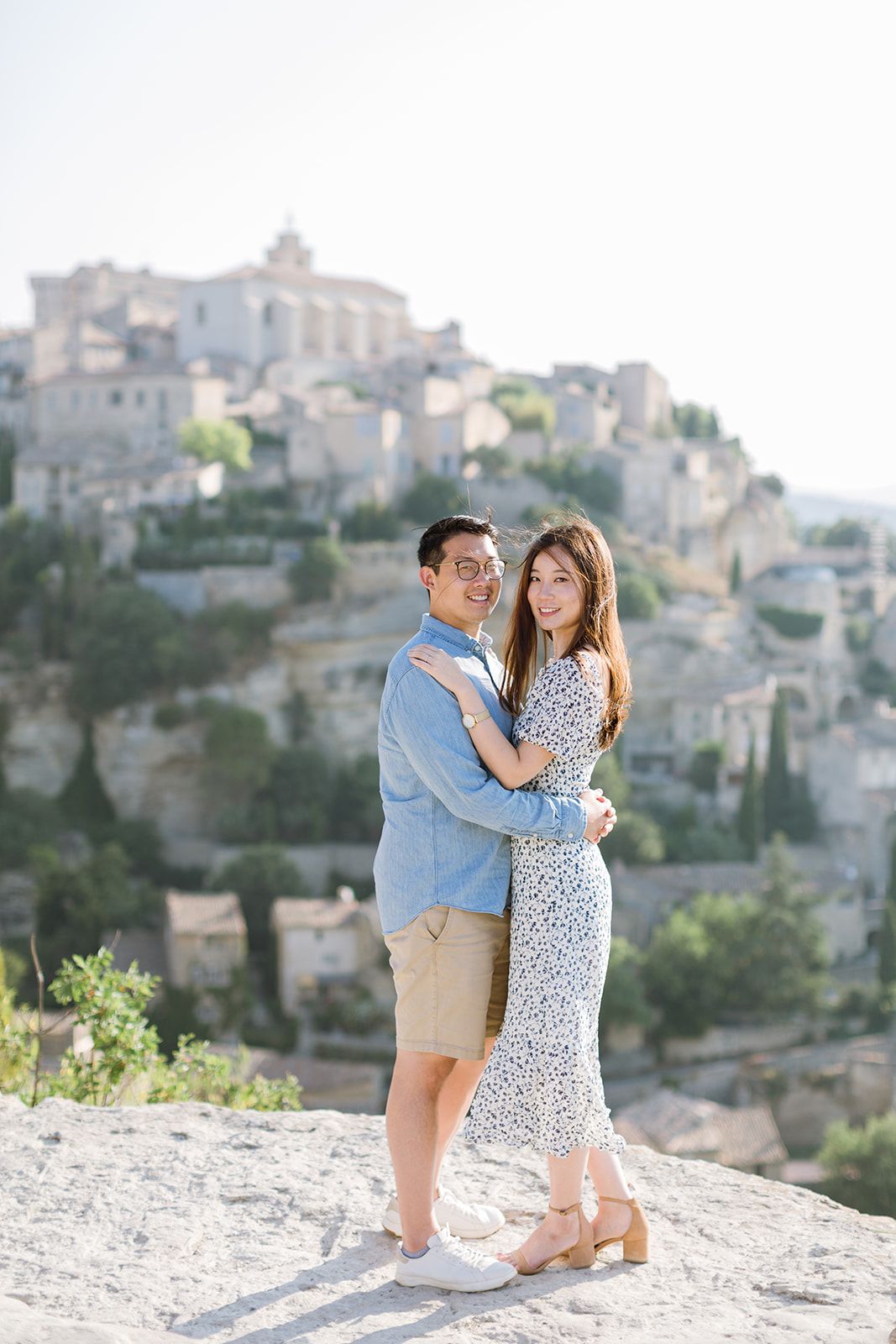 Un couple souriant s'enlace sur un promontoire rocheux, avec le village historique perché de Gordes, en France, en arrière-plan.