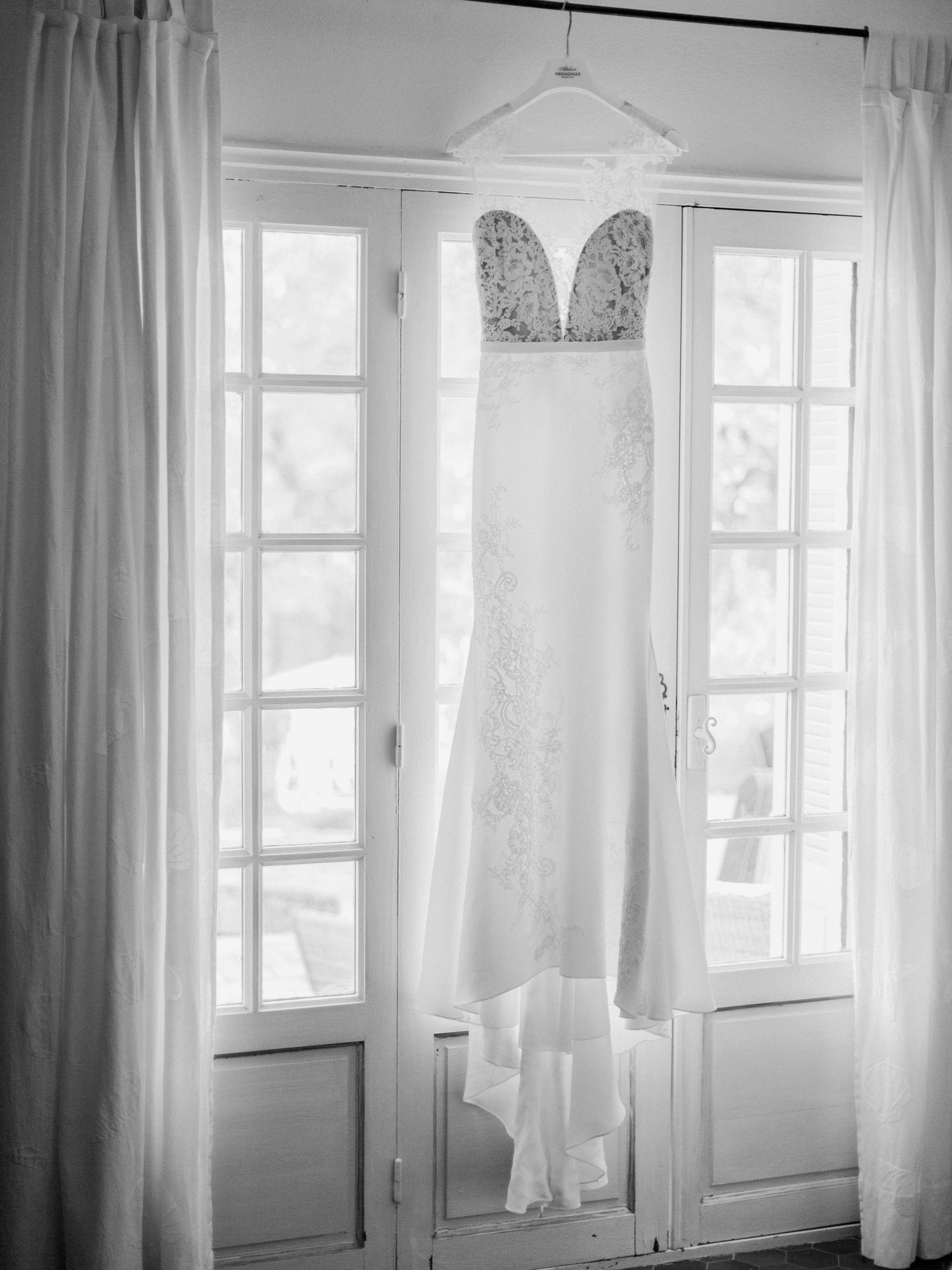 Photo en noir et blanc d'une élégante robe de mariée bustier suspendue à des portes-fenêtres avec des rideaux blancs.