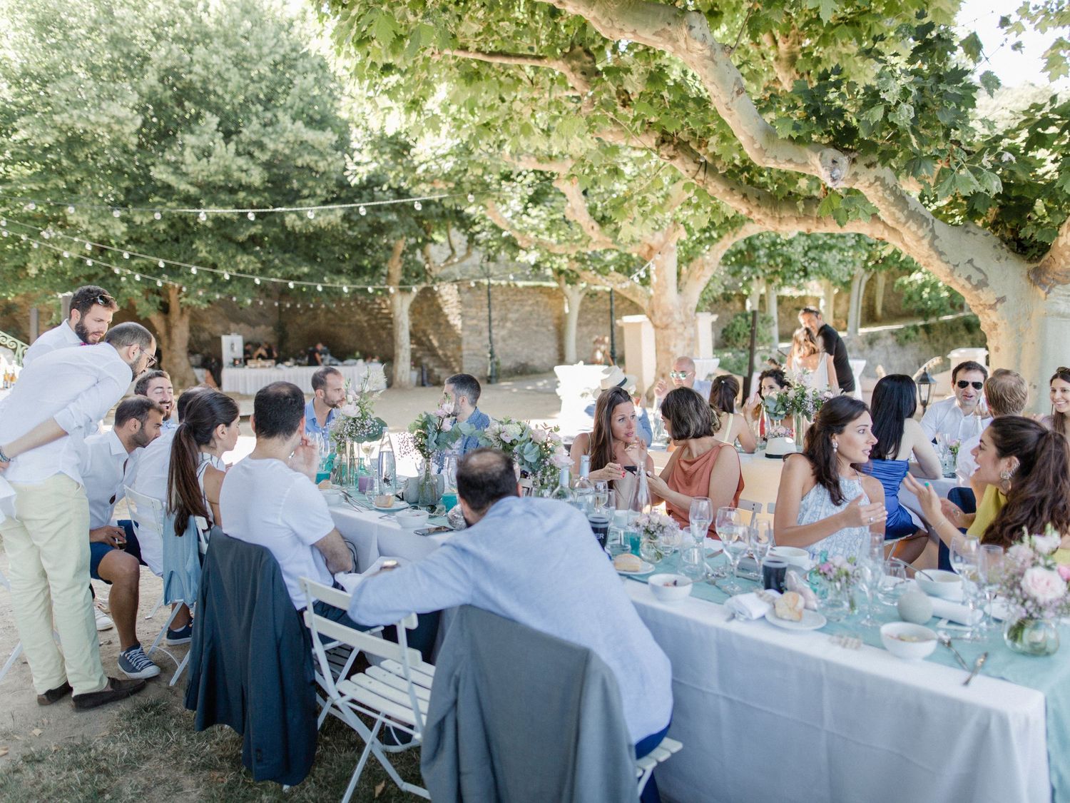 Guests gathered around a long outdoor dining table under trees with string lights at an elegant garden wedding reception.