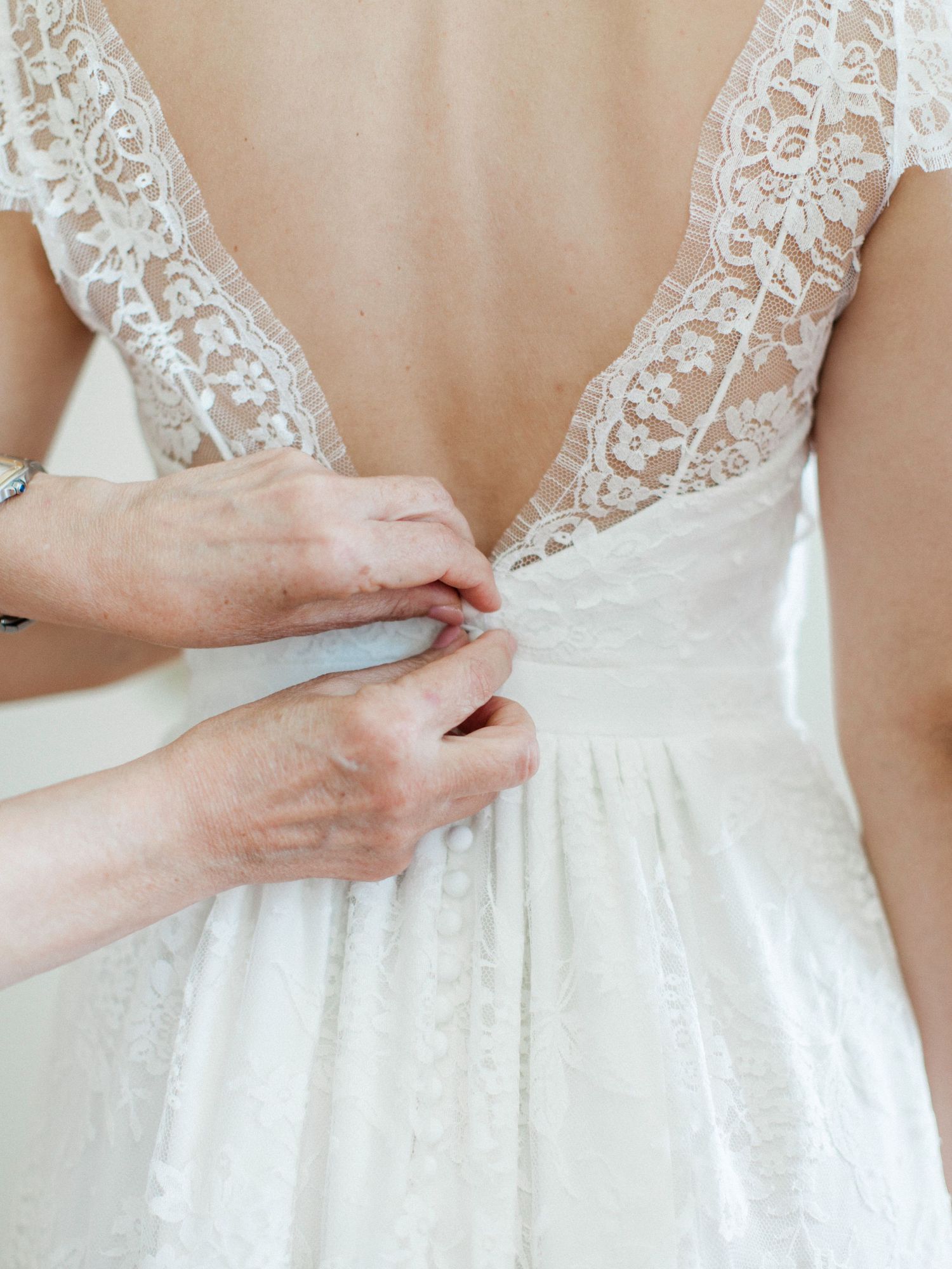 Close-up of hands fastening buttons on a white lace wedding dress with a deep V-back neckline.