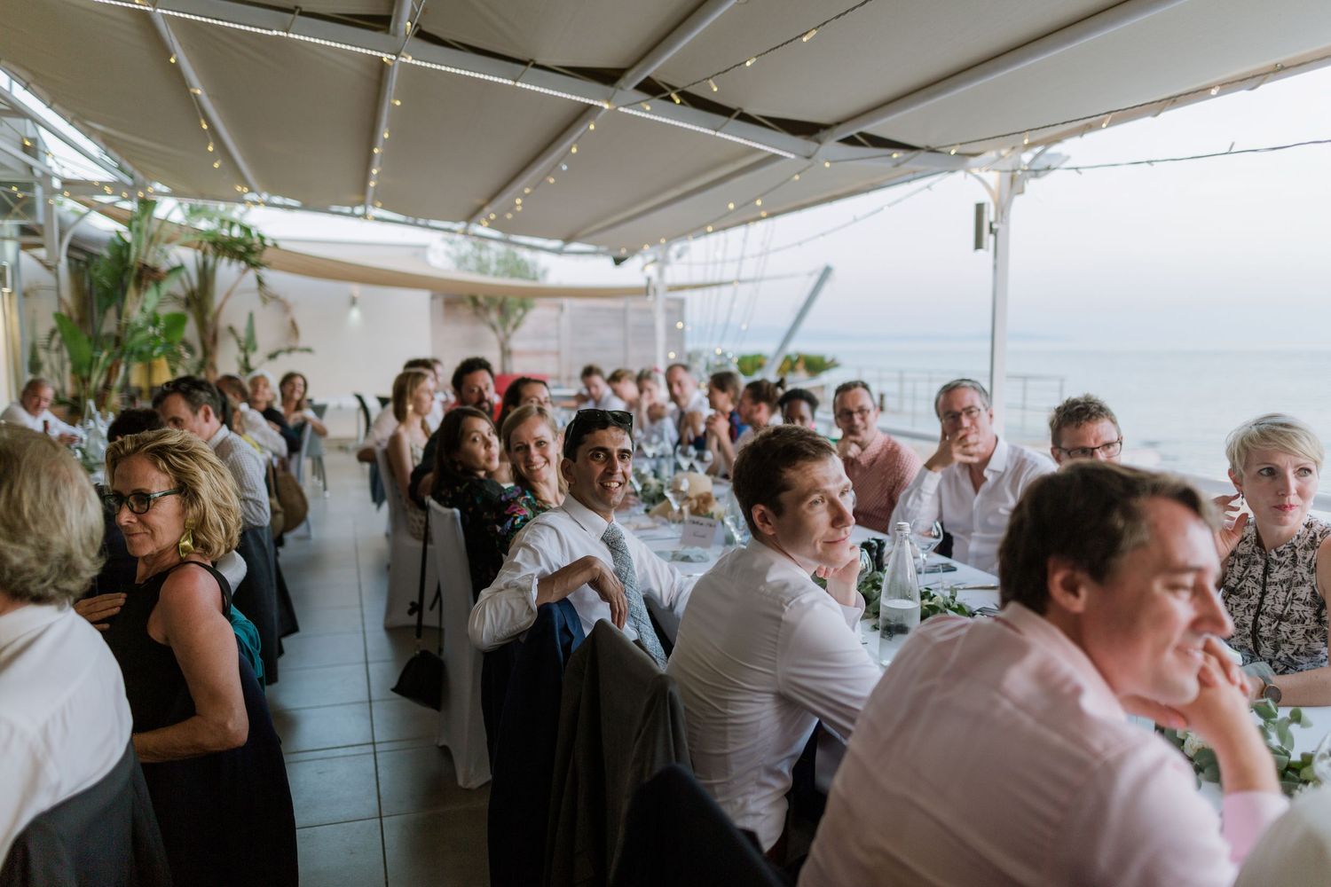 Des invités attablés lors d'une réception de mariage en plein air avec vue sur l'océan, sous une structure couverte agrémentée de guirlandes