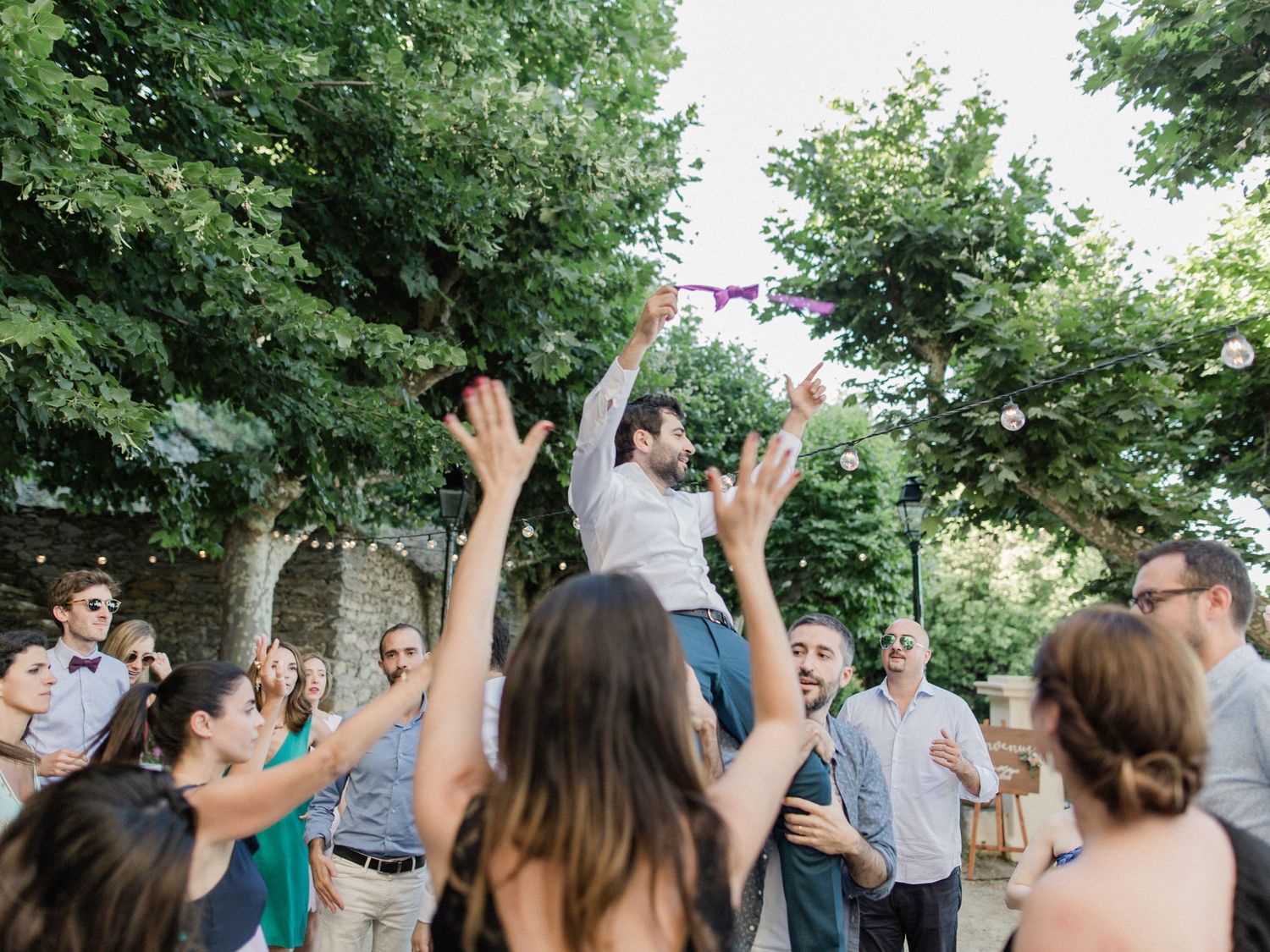 Guests celebrate outdoors at a wedding reception, with a man lifted on shoulders tossing a bouquet among cheering crowd.