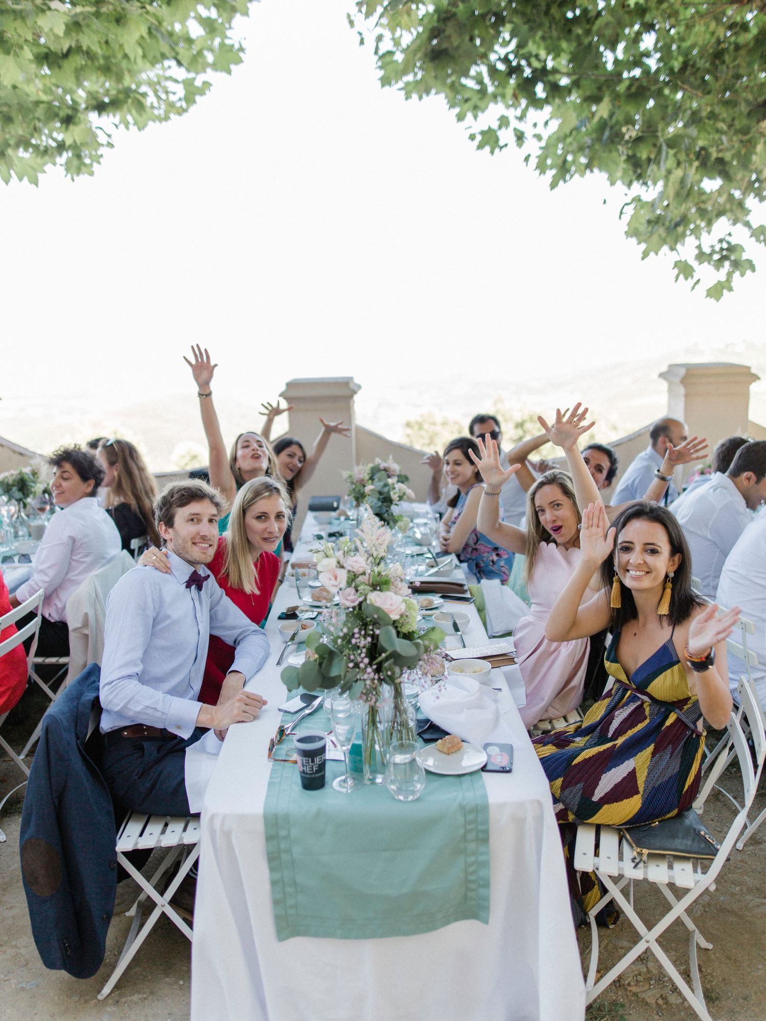 Guests celebrate at an outdoor wedding reception, raising hands joyfully at a long decorated table surrounded by trees.