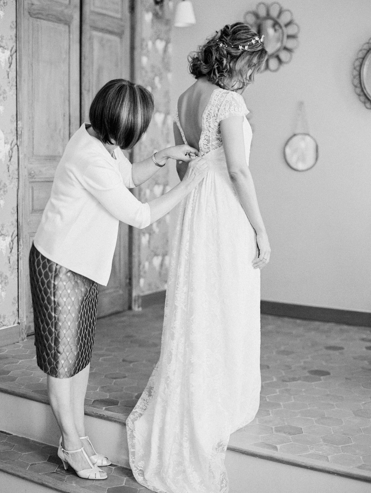 Black and white photo of a woman helping a bride button her lace wedding dress before the ceremony.
