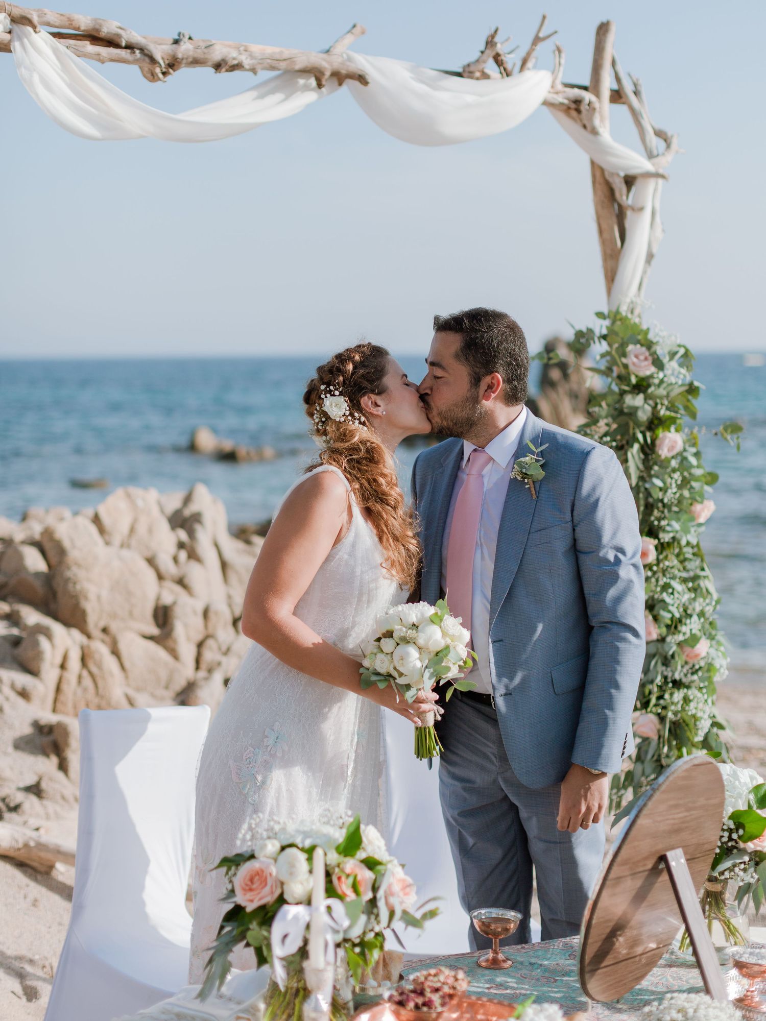 Les mariés échangent un baiser lors d'une cérémonie romantique sur la plage, sous une arche en bois flotté agrémentée de décorations florale
