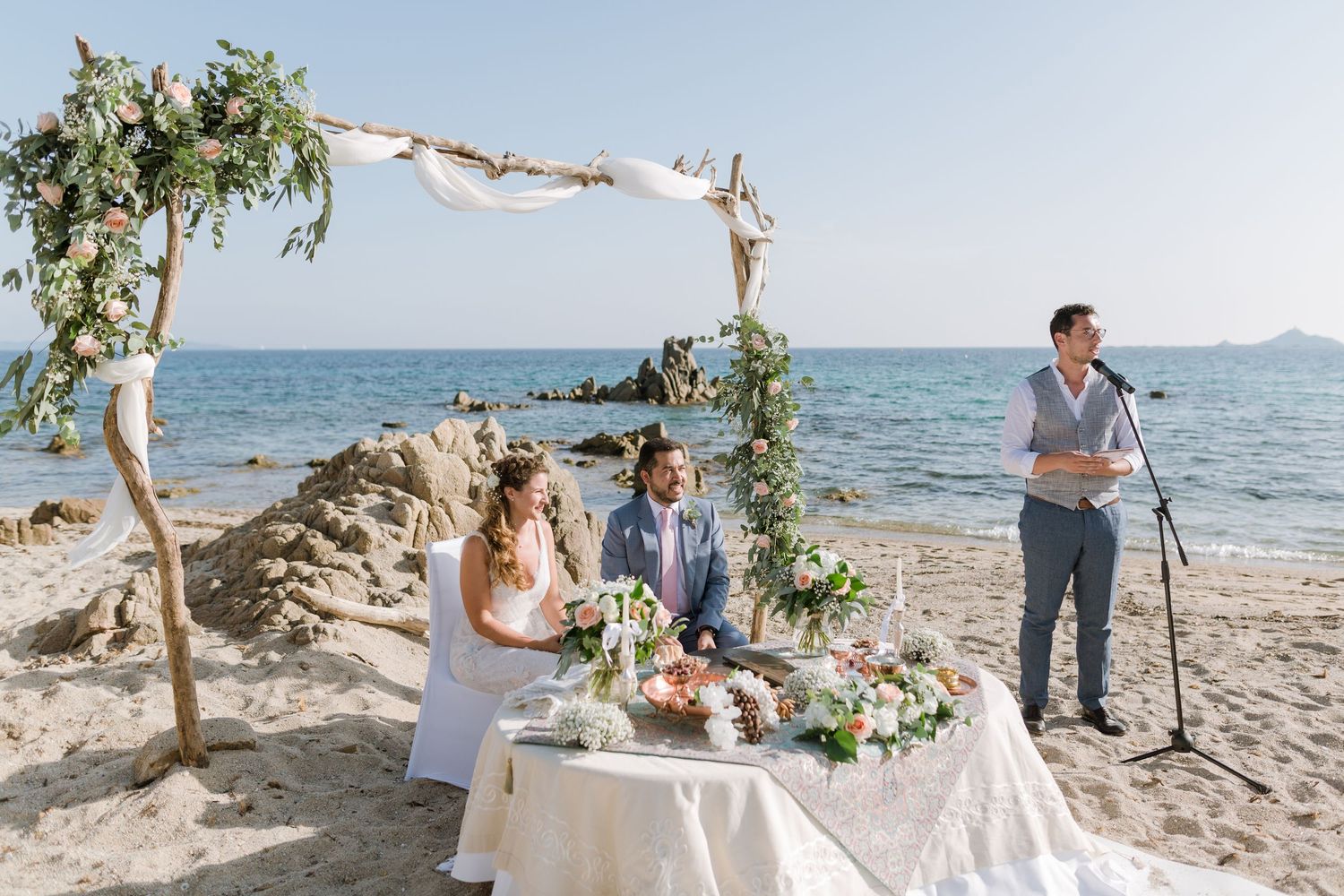 Un couple échange ses vœux lors d'une cérémonie de mariage romantique sur la plage, sous une arche fleurie avec vue sur l'océan.