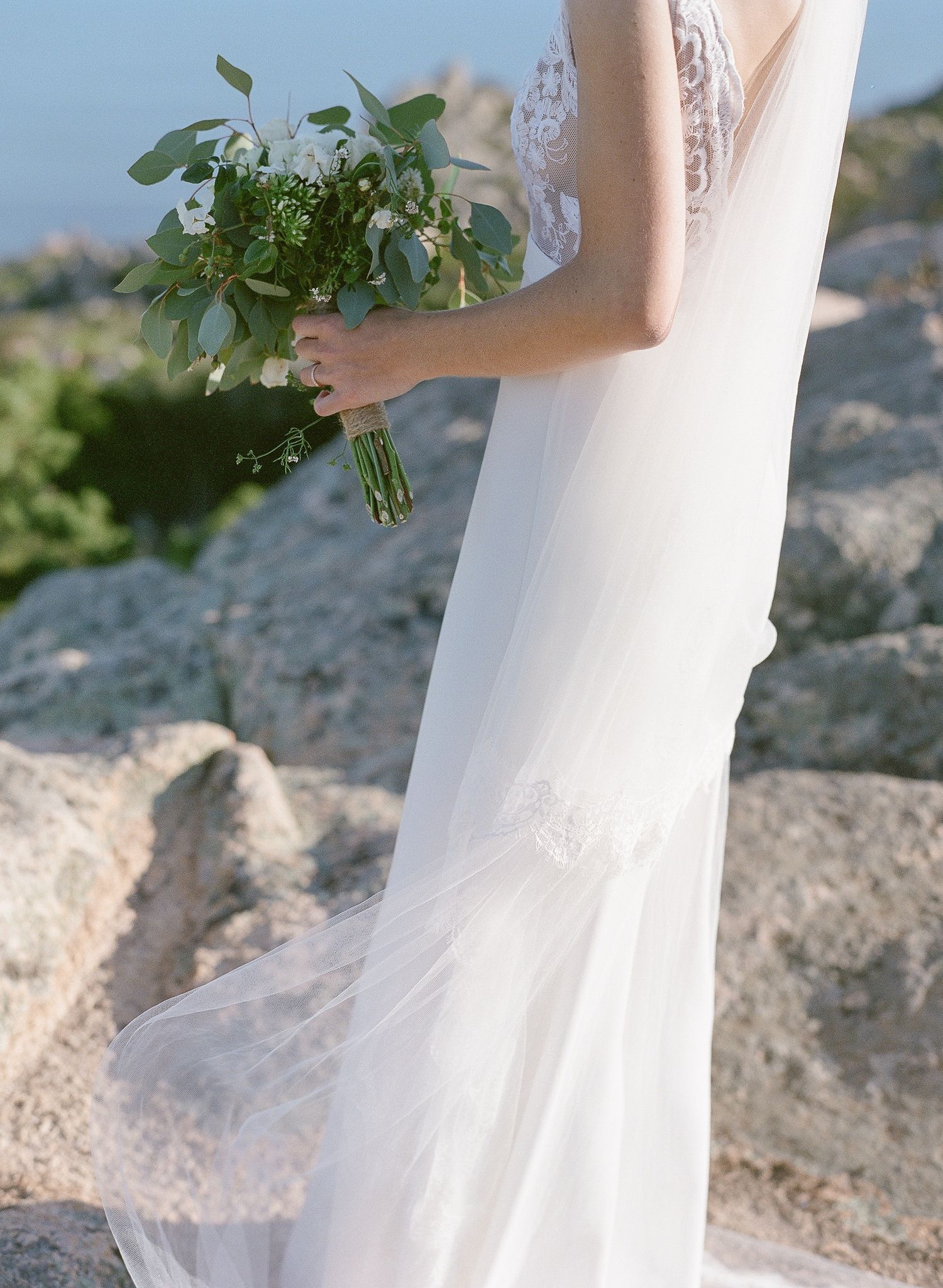 Une mariée en élégante robe de mariée blanche tient un bouquet vert et blanc sur un terrain rocailleux en extérieur.