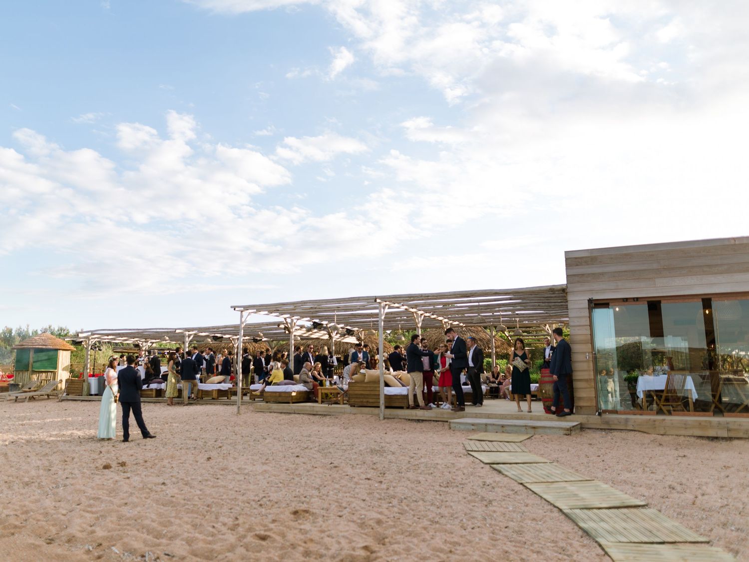 Réception de mariage en plein air sur une plage de sable fin, les invités se mêlant sous une pergola moderne par une journée ensoleillée.