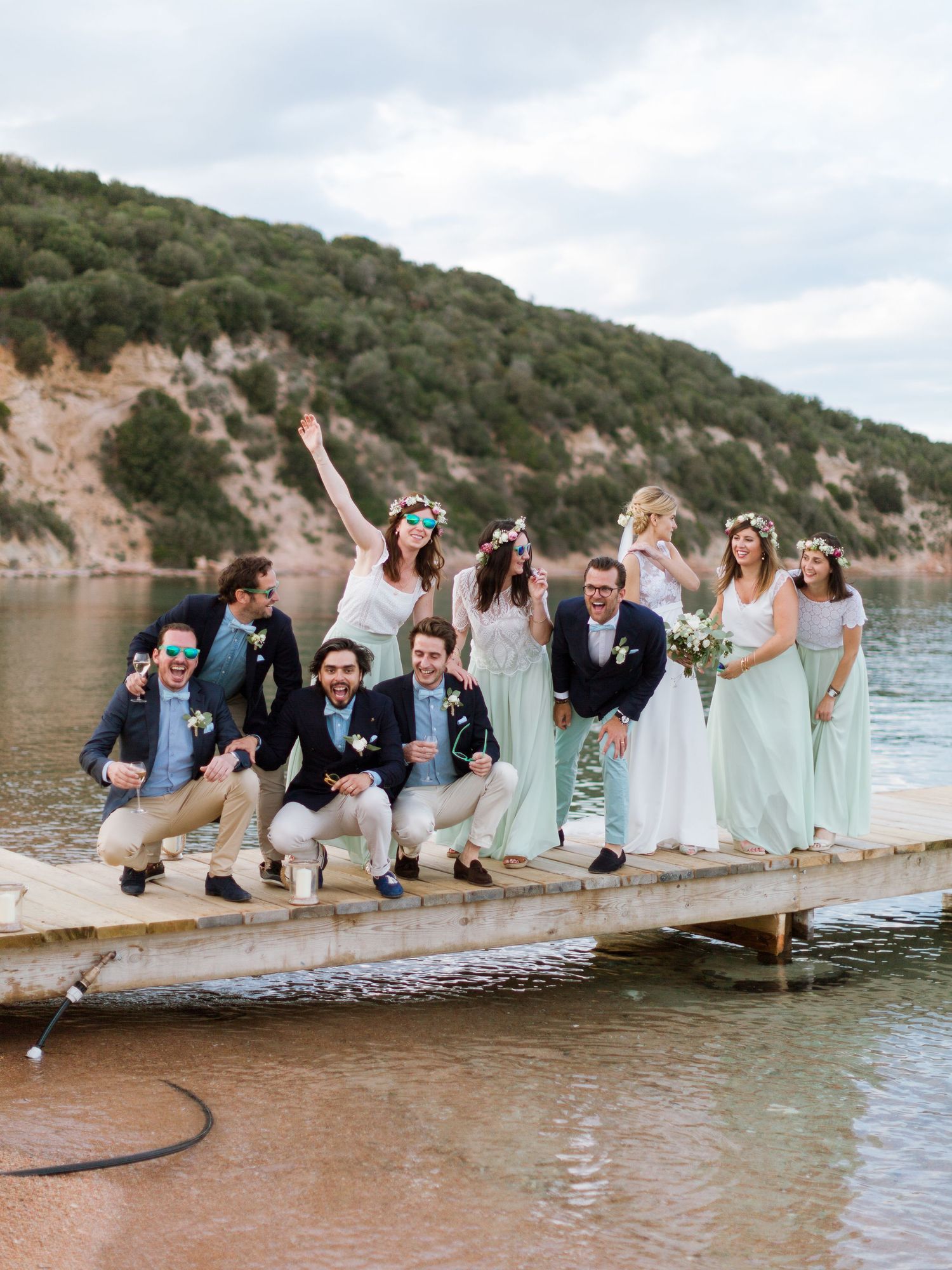Le cortège nuptial pose joyeusement sur un quai au bord d'un lac, les demoiselles d'honneur en robes menthe avec des couronnes de fleurs