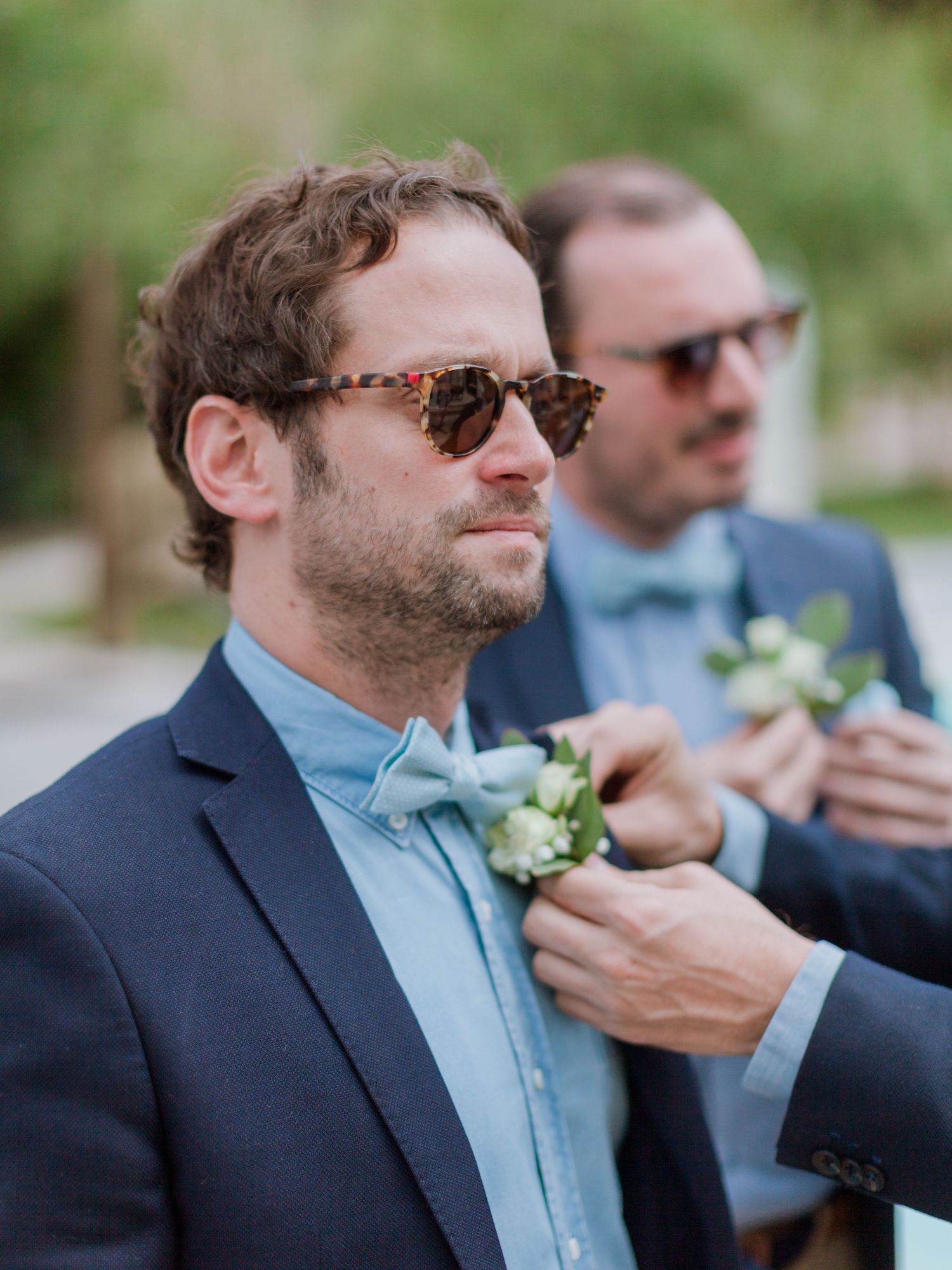 Des garçons d'honneur en costumes bleu marine et nœuds papillon se font épingler des boutonnières, portant des lunettes de soleil