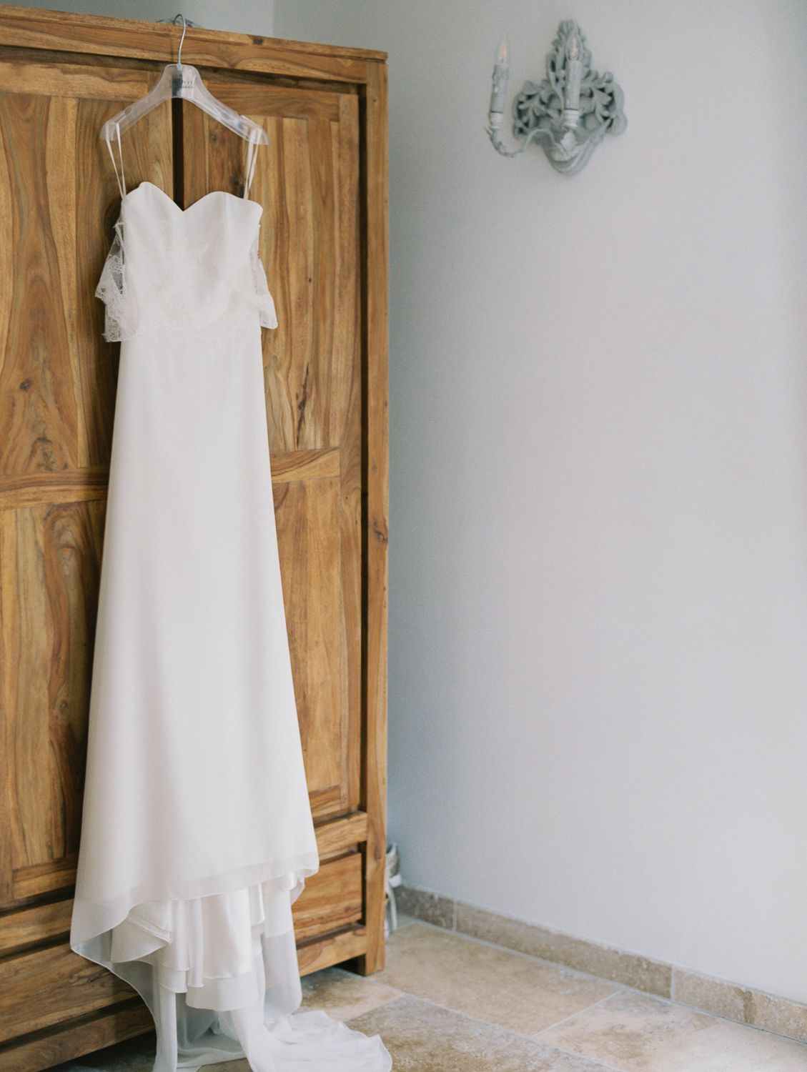 Elegant white strapless wedding gown with flowing train hanging on a wooden armoire in a bright room.