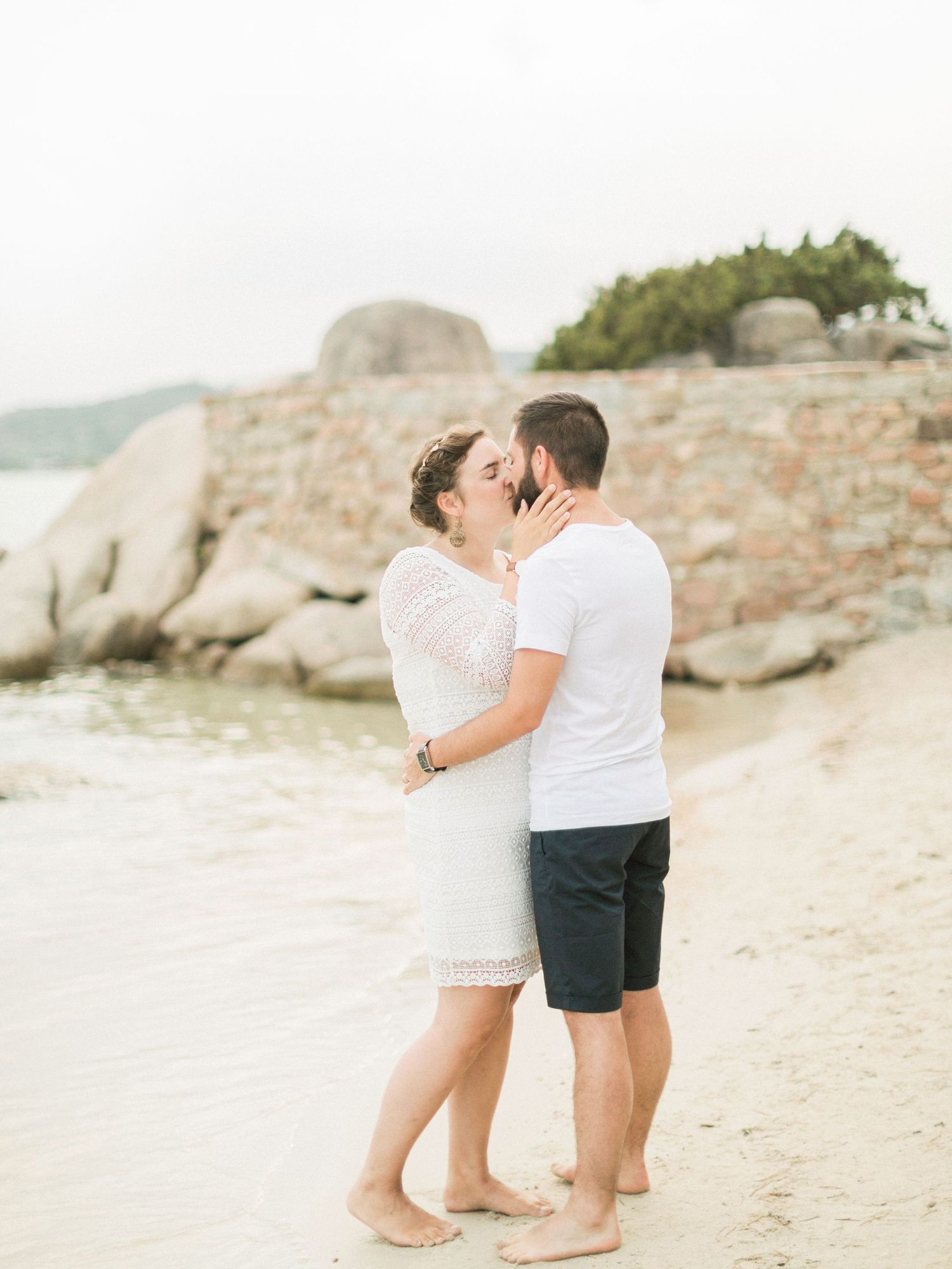 Un couple échange un baiser romantique sur une plage de sable fin, près d'anciennes ruines de pierre au bord de l'eau.