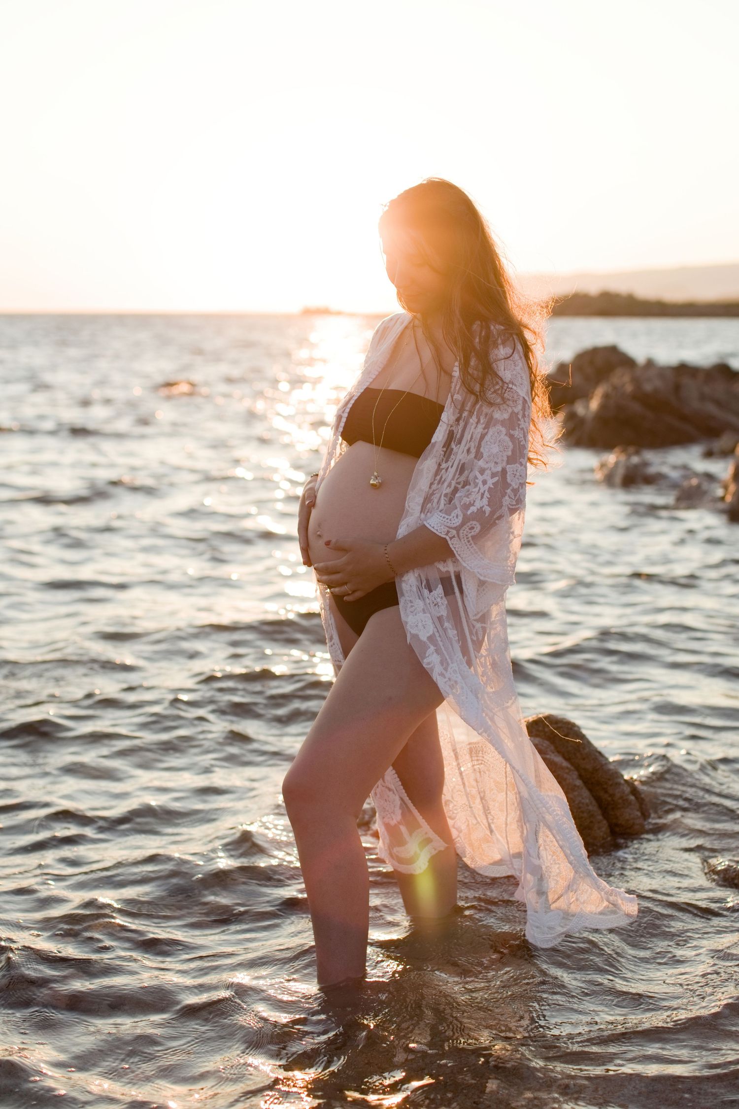 Femme enceinte vêtue d'un paréo en dentelle, debout dans les eaux peu profondes de la mer  au coucher du soleil près de Bonifacio.