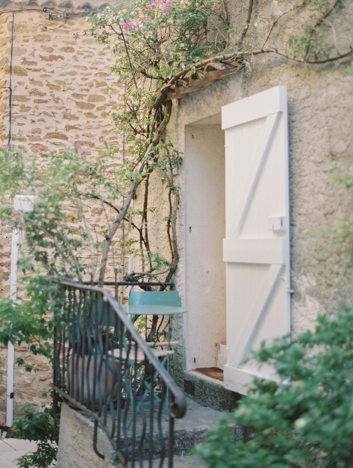 Bâtiment rustique en pierre avec porte à volets blancs, rosiers grimpants et balustrade en fer forgé, dans un charmant village français