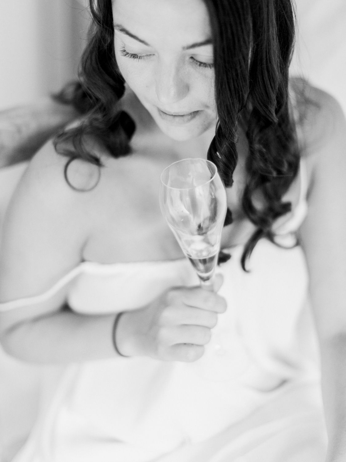 Photo en noir et blanc d'une mariée tenant une flûte de champagne, le regard baissé et arborant un doux sourire le jour de son mariage.