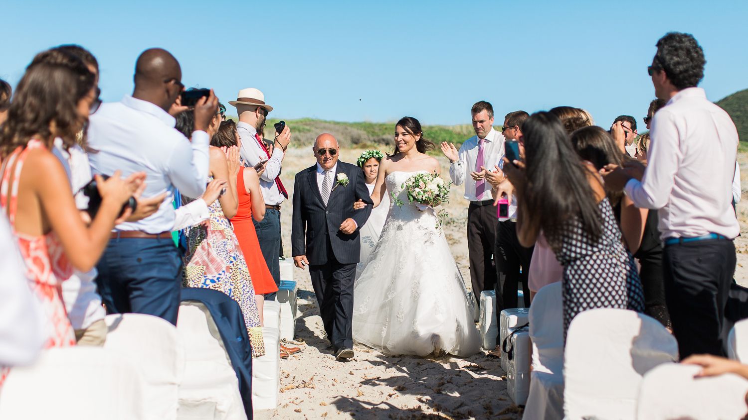 La mariée remonte l'allée de sable fin, accompagnée de son escorte, entourée d'invités souriants lors d'une cérémonie de mariage