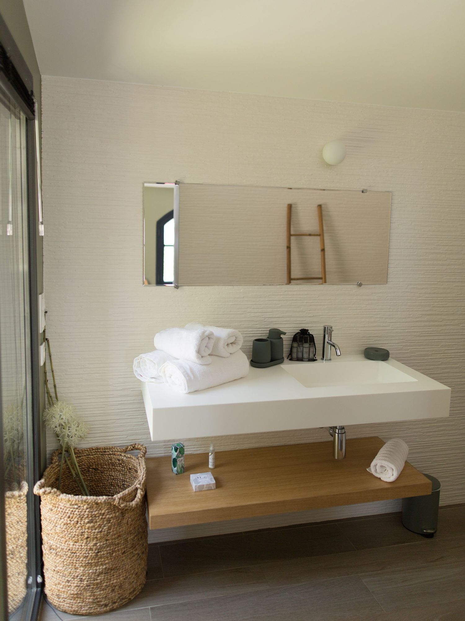 Salle de bains minimaliste blanche avec lavabo suspendu, étagère en bois, panier en osier et décoration murale à motifs botaniques.