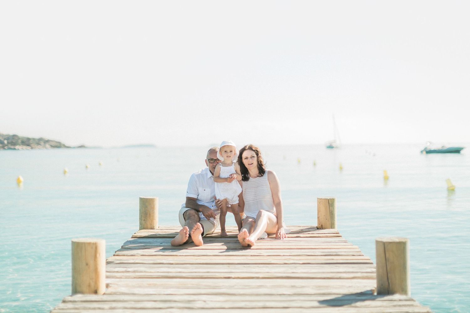 Une famille souriante avec un jeune enfant pose sur un quai en bois entouré d'une eau d'un bleu limpide. Sur la plage de Santa giulia.