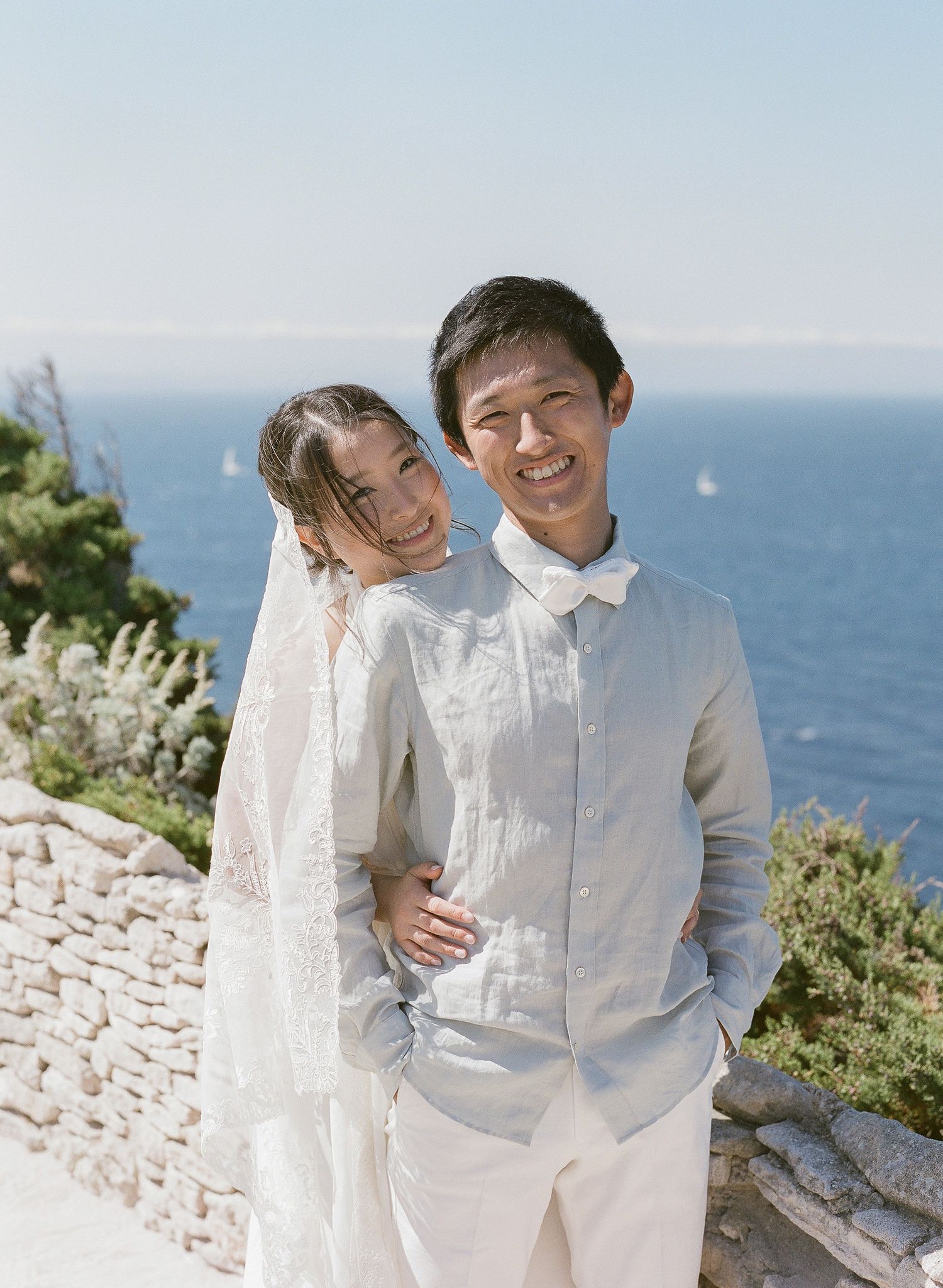 Un couple asiatique en tenue de mariage sourit et s'enlace devant un magnifique fond d'océan bleu par une journée ensoleillée à Bonifacio