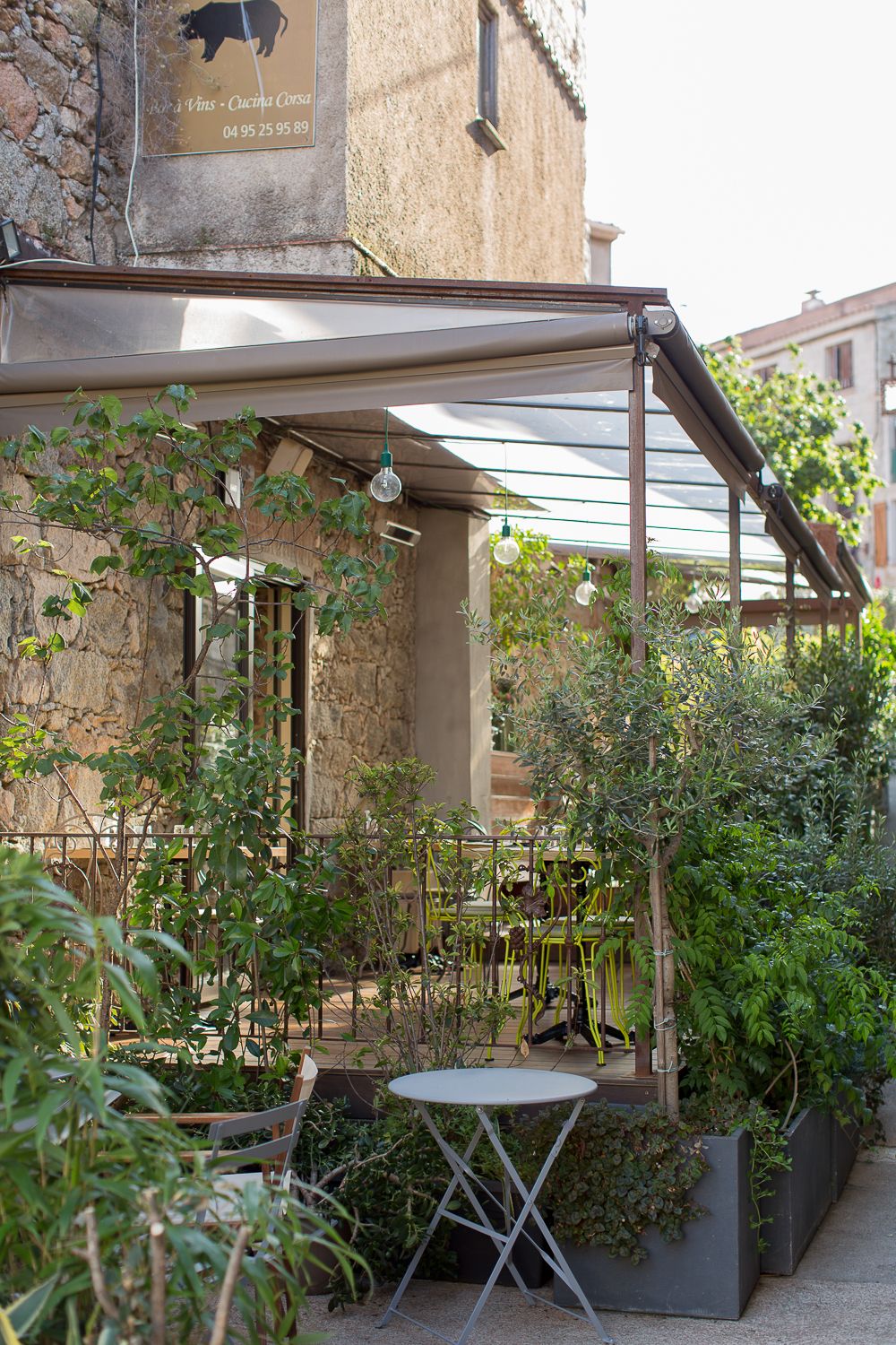 Façade en pierre d'un bâtiment avec terrasse de restaurant couverte, plantes luxuriantes et petite table et chaise de bistro à L'orriu.