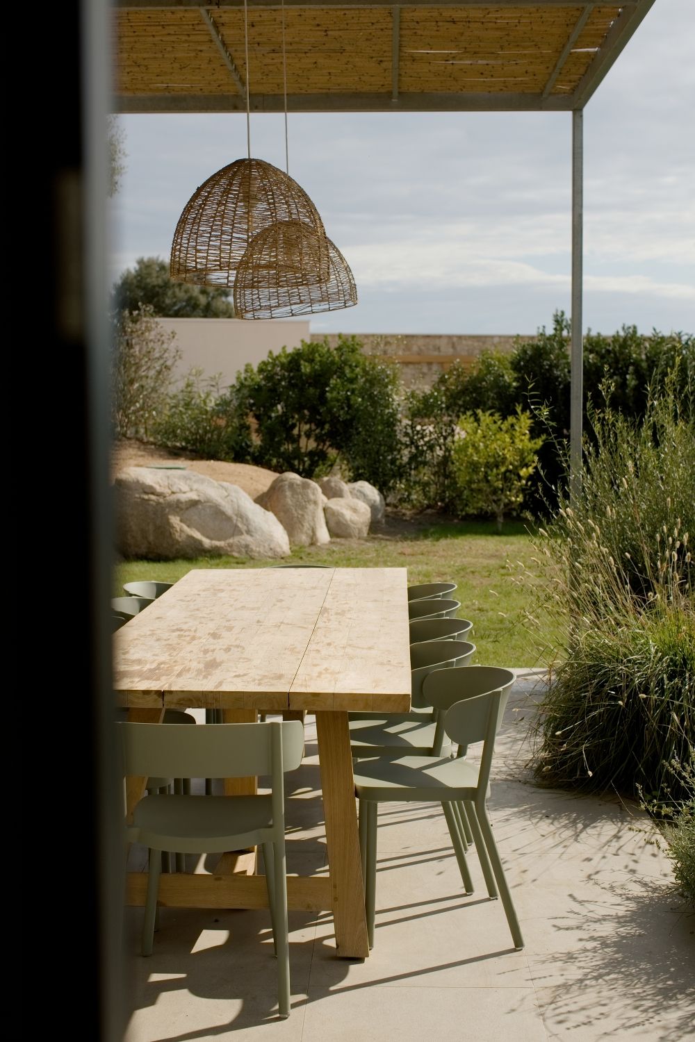 Coin repas extérieur avec table rustique en bois, chaises vert sauge, suspension lumineuse en osier et jardin au-delà.