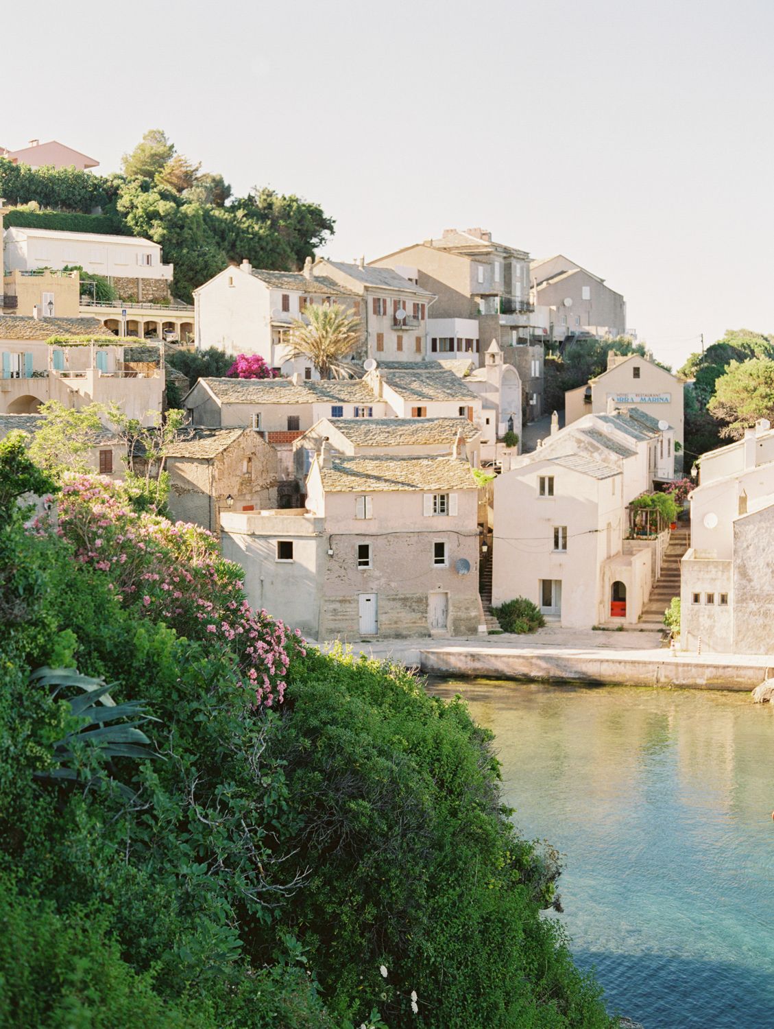 Village côtier pittoresque avec ses bâtiments en pierre, sa végétation luxuriante et son eau turquoise cristalline.