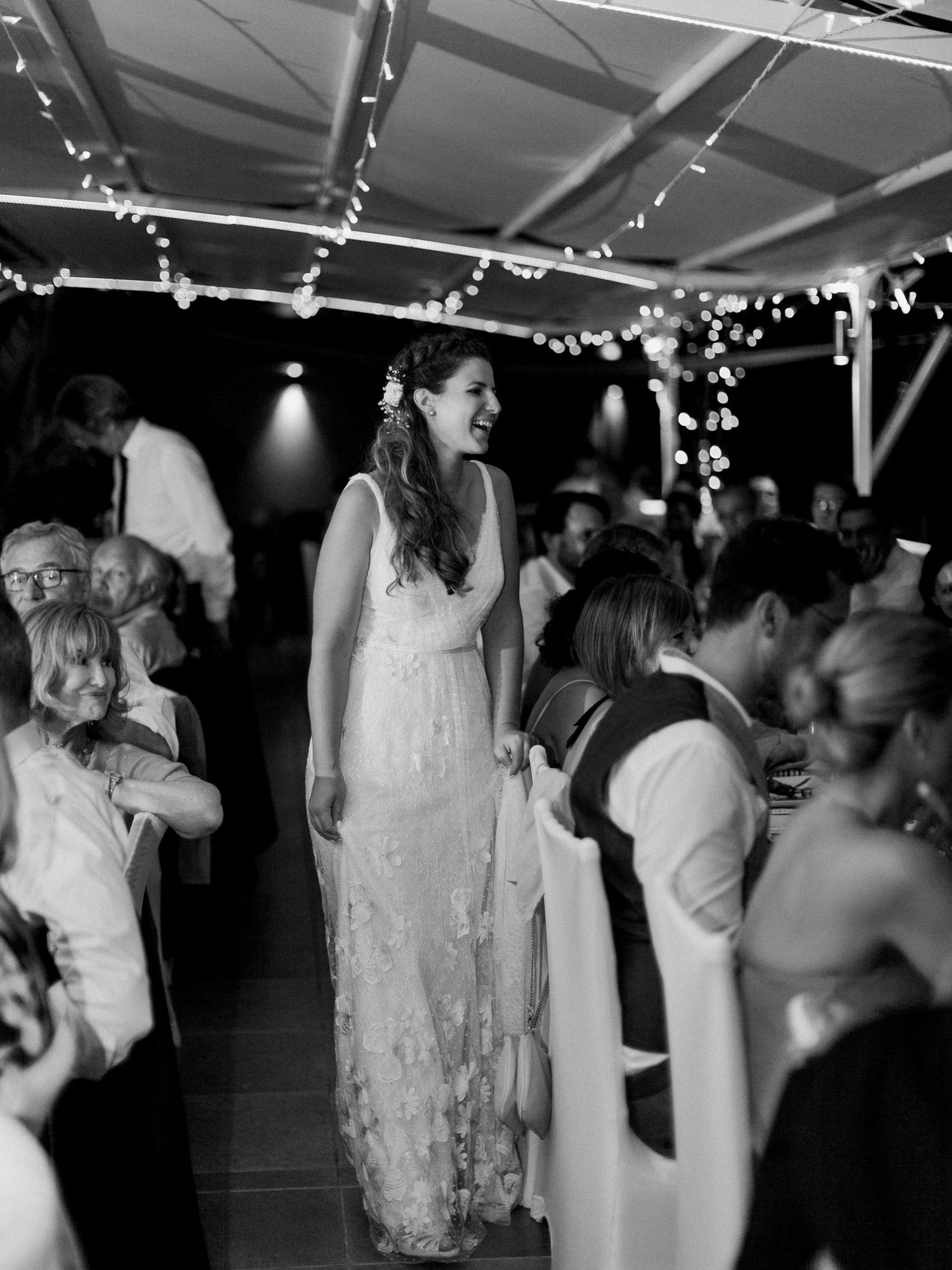 Une mariée en robe de dentelle rit joyeusement lors d'une réception en plein air en soirée, illuminée par des guirlandes lumineuses.