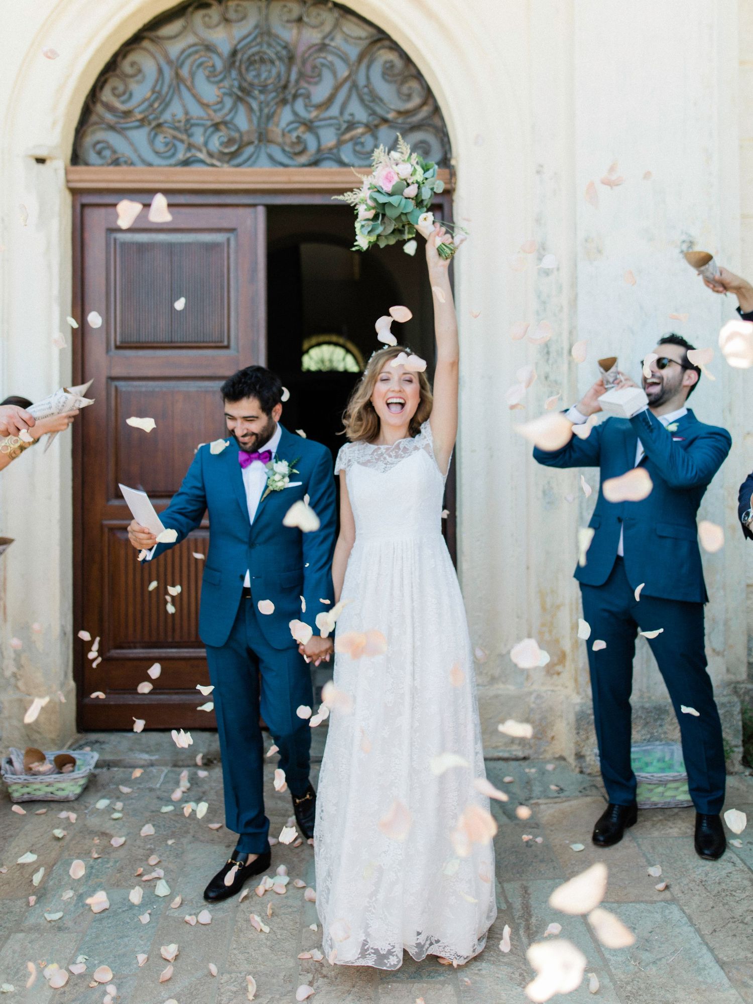 Joyful bride and groom exit church doors as wedding guests toss flower petals in celebration.