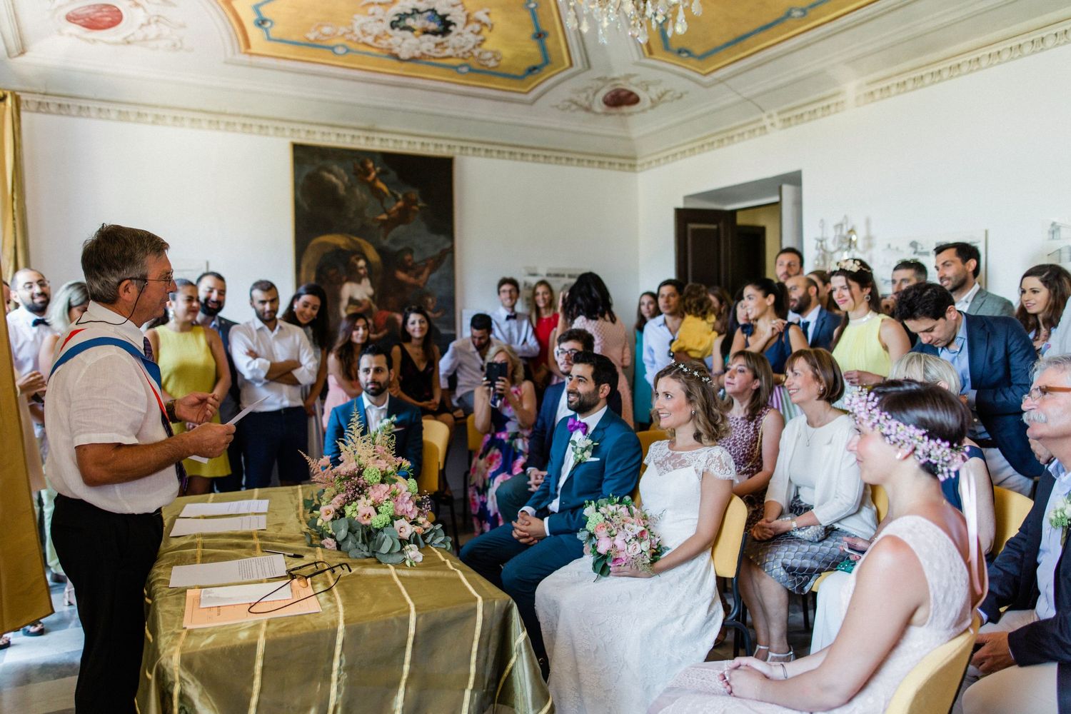 Wedding ceremony in an ornate room with guests watching as officiant addresses smiling couple seated at decorated table.