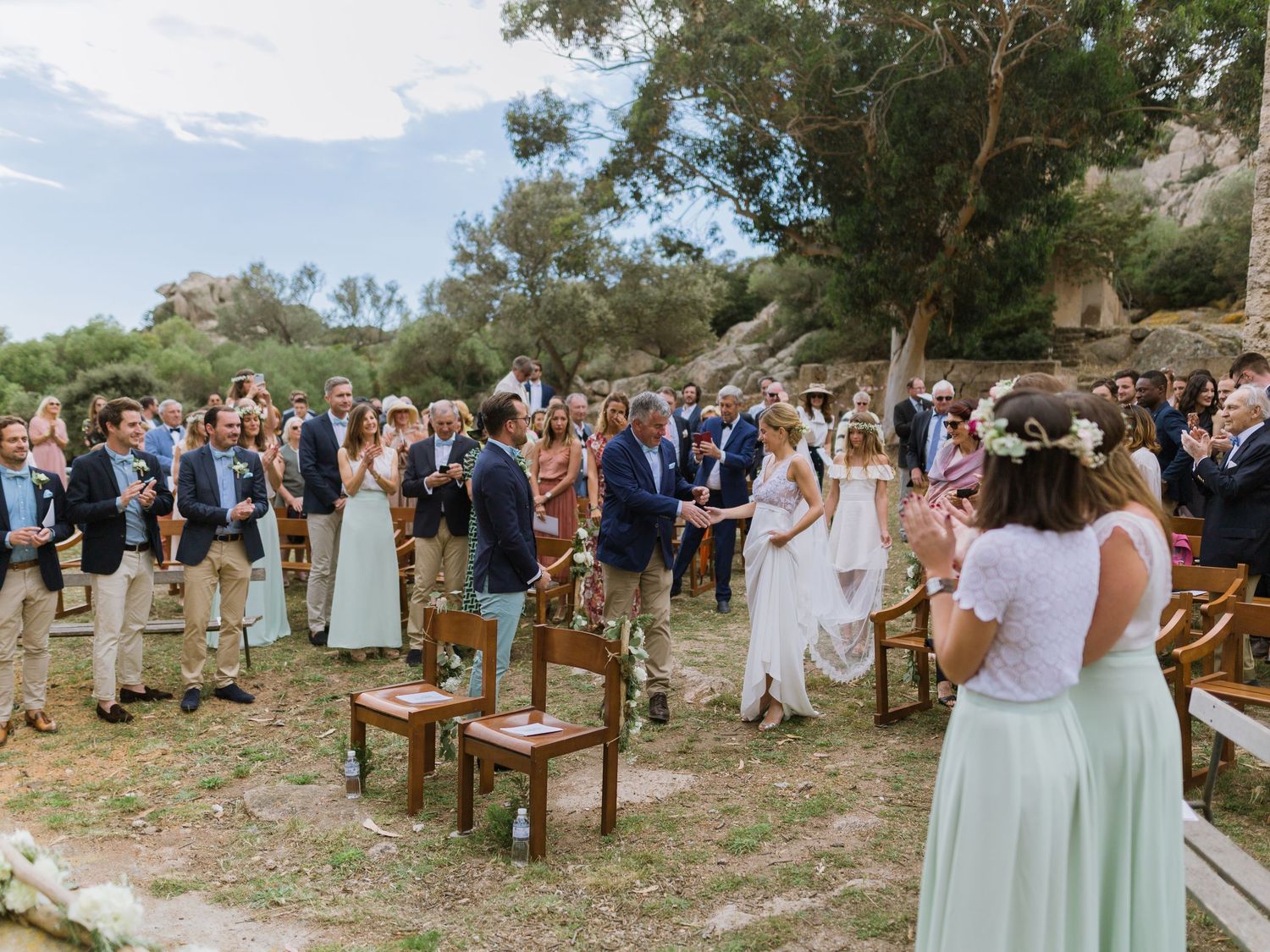 Cérémonie de mariage en plein air, les invités rassemblés parmi les arbres, la mariée en robe blanche remonte l'allée dans un cadre naturel