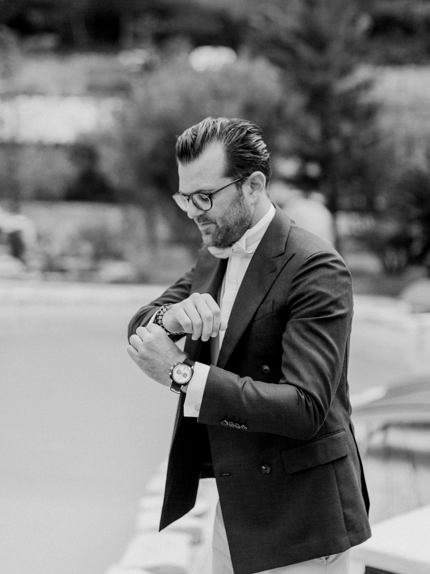 Un homme élégant en costume sombre ajuste sa montre en regardant vers le bas, debout à l'extérieur sur une photo en noir et blanc.