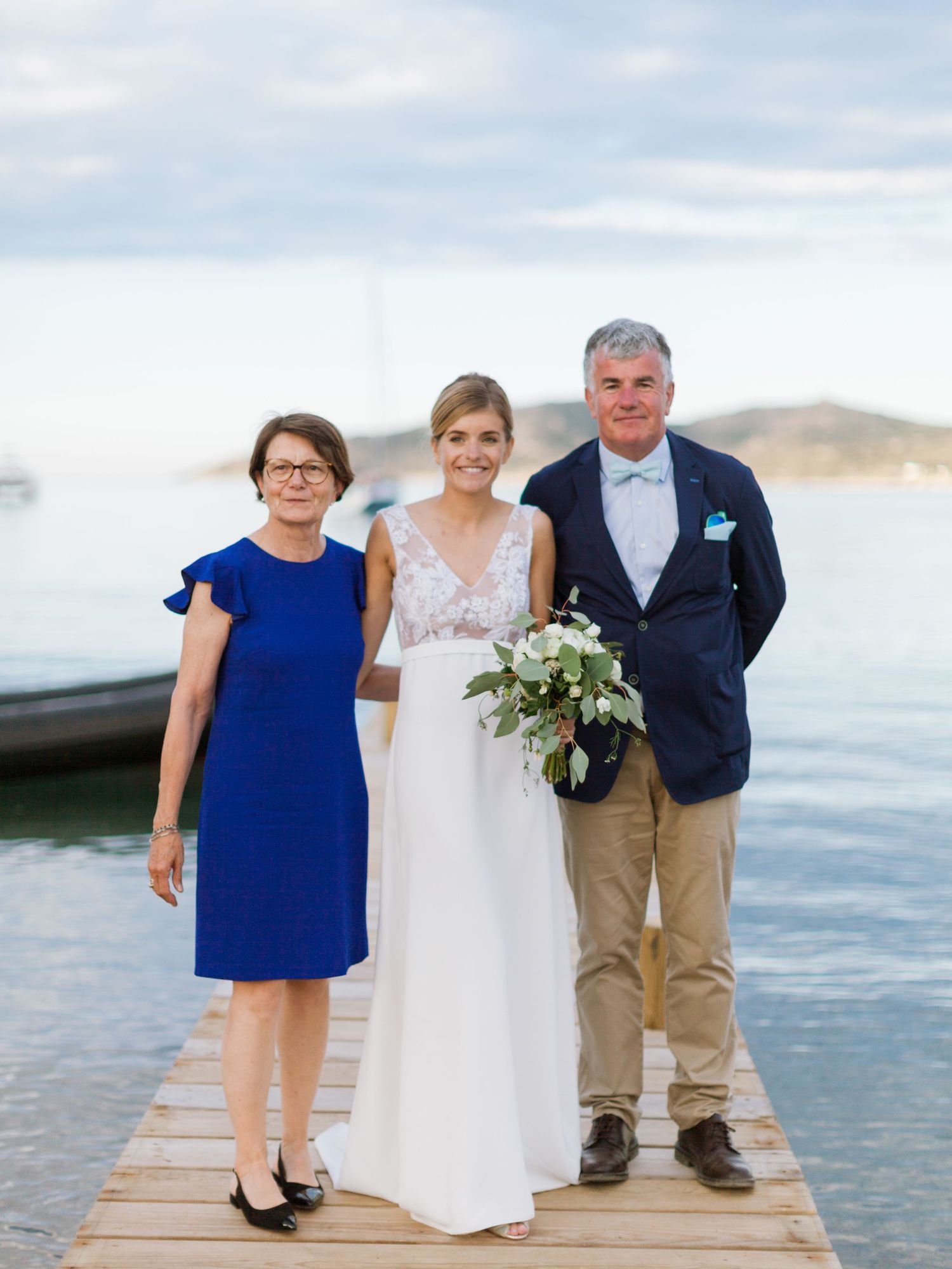 La mariée en robe blanche tient son bouquet, entourée de ses parents, sur un quai avec vue sur l'océan lors d'un mariage en bord de mer.