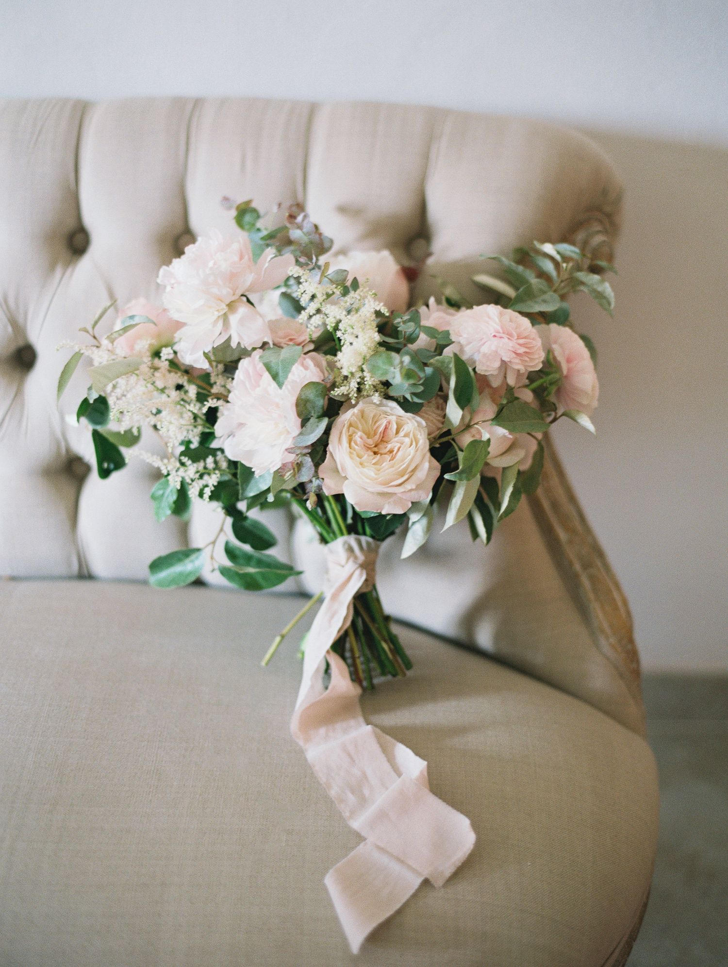 Bouquet de mariée romantique composé de roses blush et de verdure, noué d'un ruban de soie, posé sur une chaise ivoire capitonnée.