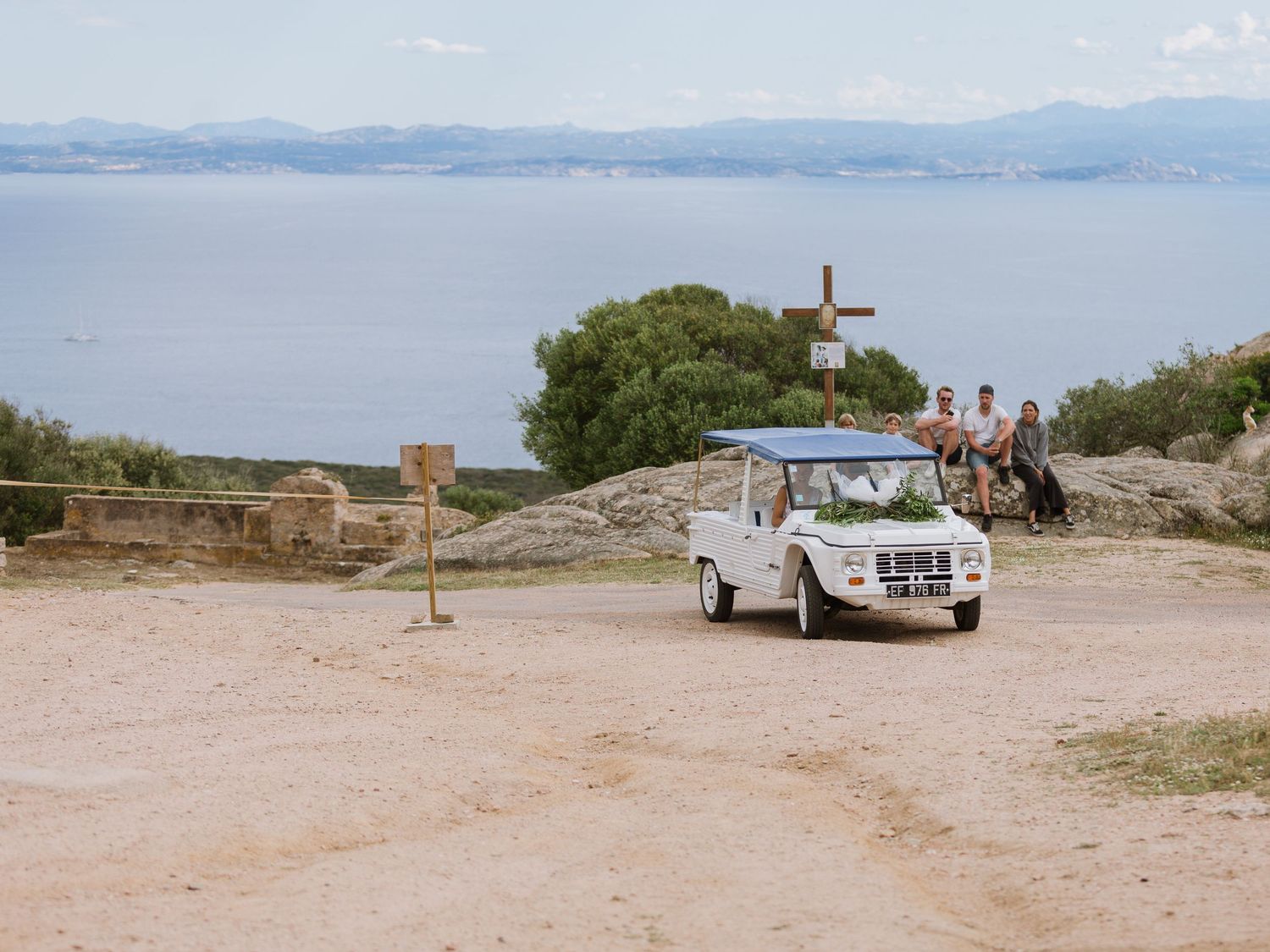 Citroën Méhari blanche vintage sur un point de vue côtier pittoresque, avec une croix en bois et une vue sur l'océan en arrière-plan.