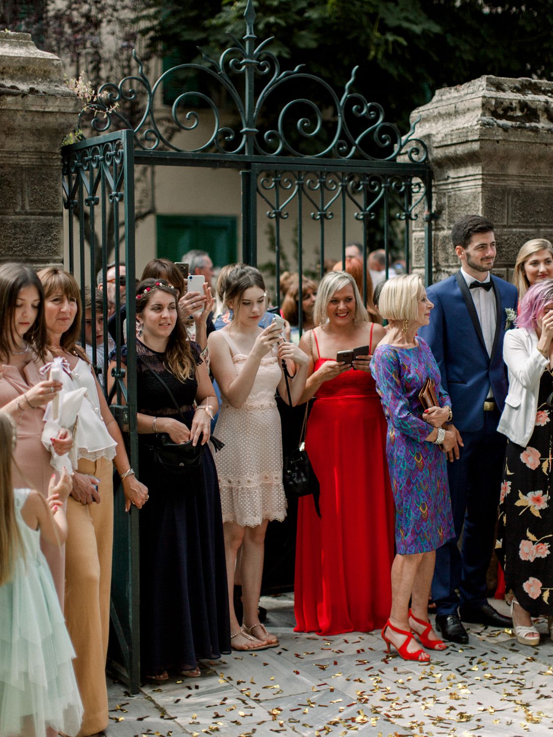 Wedding guests gathered by ornate iron gates, dressed in formal attire, celebrating outdoors on a sunny day.