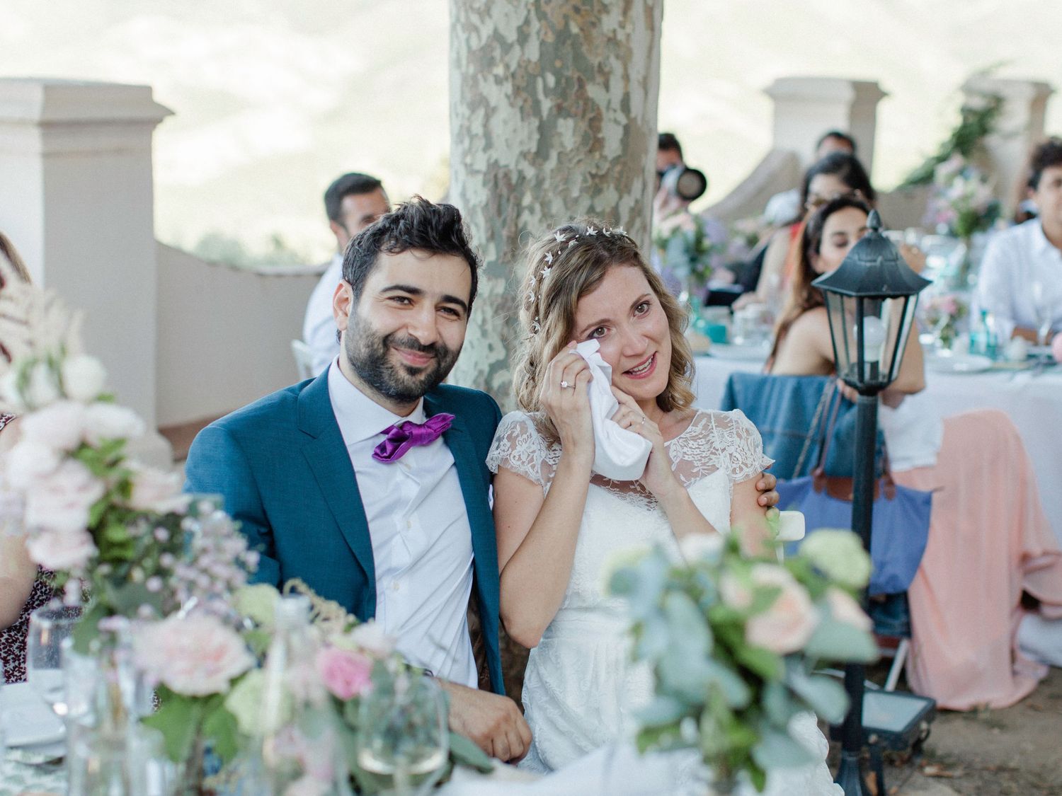 A bride and groom smile at their outdoor wedding reception, surrounded by floral arrangements and guests.