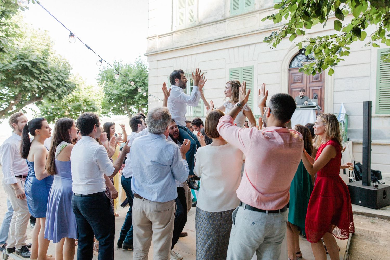Guests celebrate outdoors at a wedding reception, clapping and dancing near a elegant building with string lights.