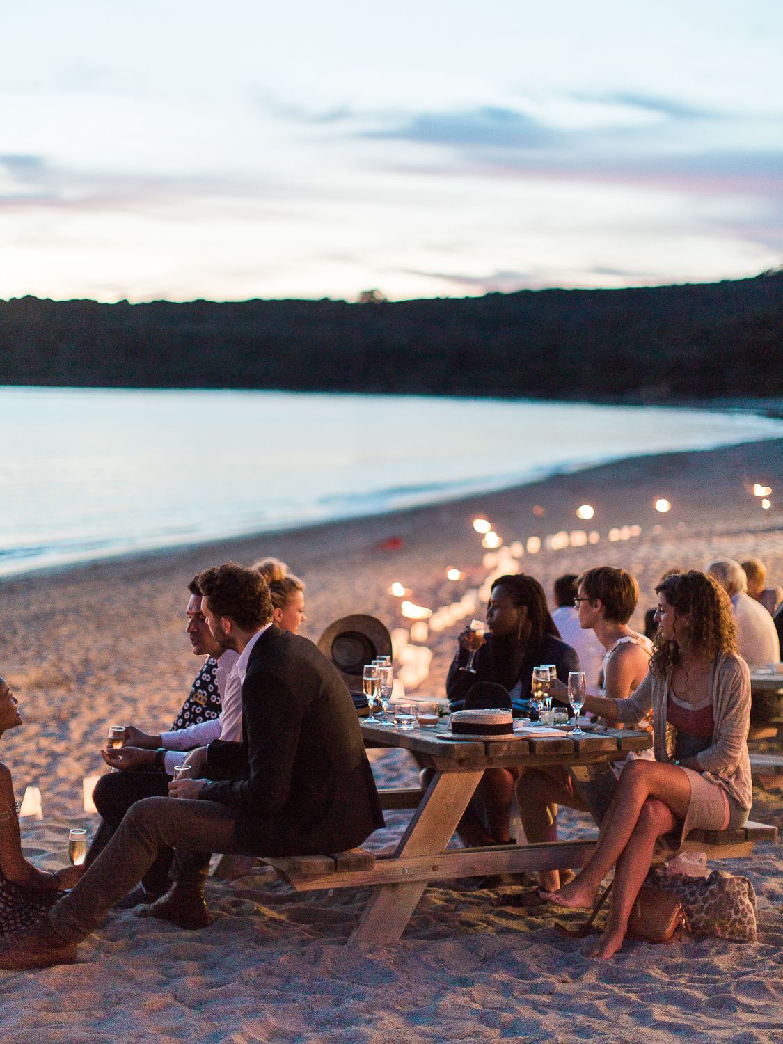 Un groupe d'amis dîne à une table de pique-nique sur une plage de sable au crépuscule, des bougies éclairant le rivage à  Ajaccio