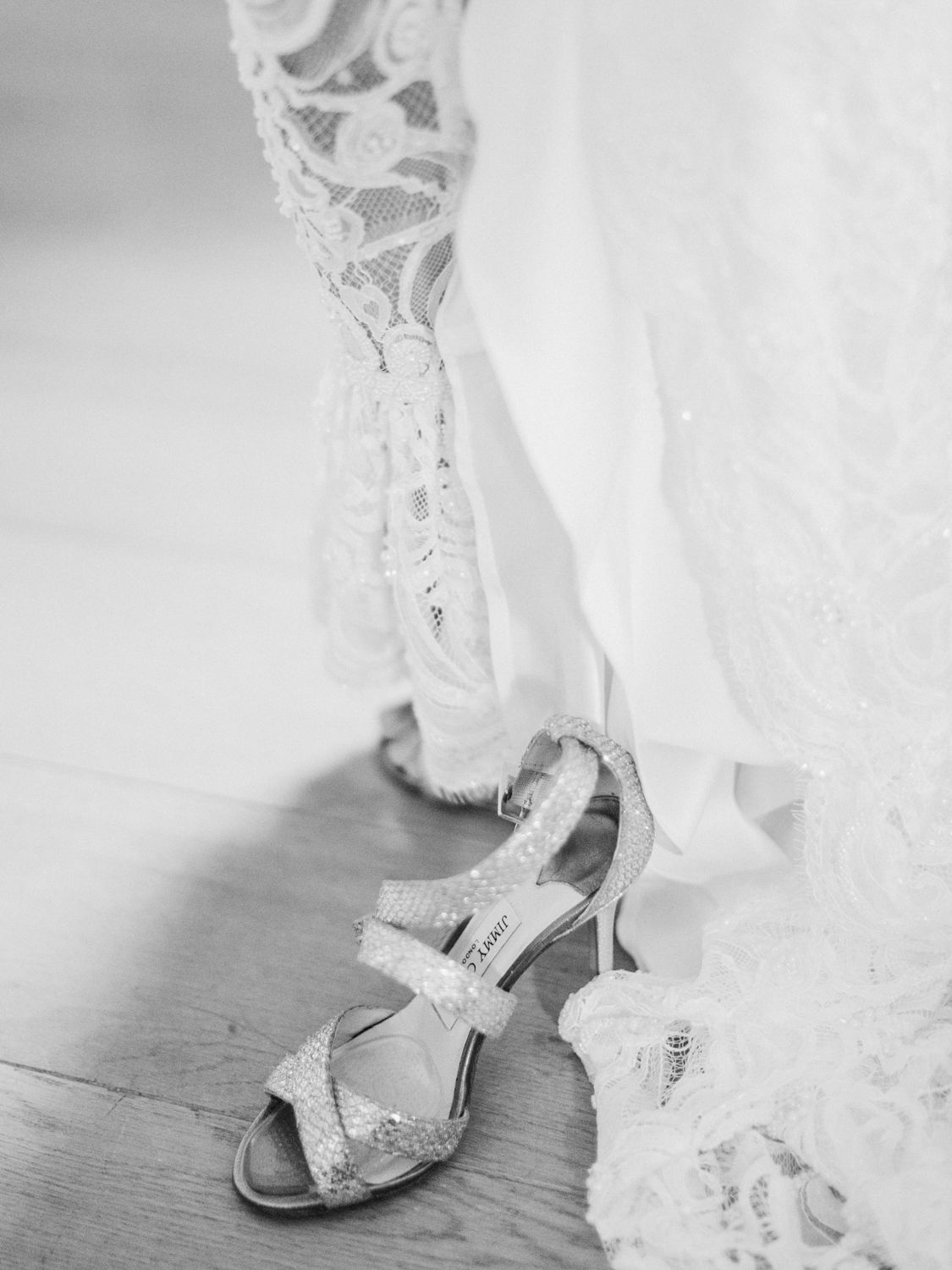 Photo en noir et blanc de talons de mariée à paillettes, posés à côté d'un ourlet de robe de mariée en dentelle sur un parquet.