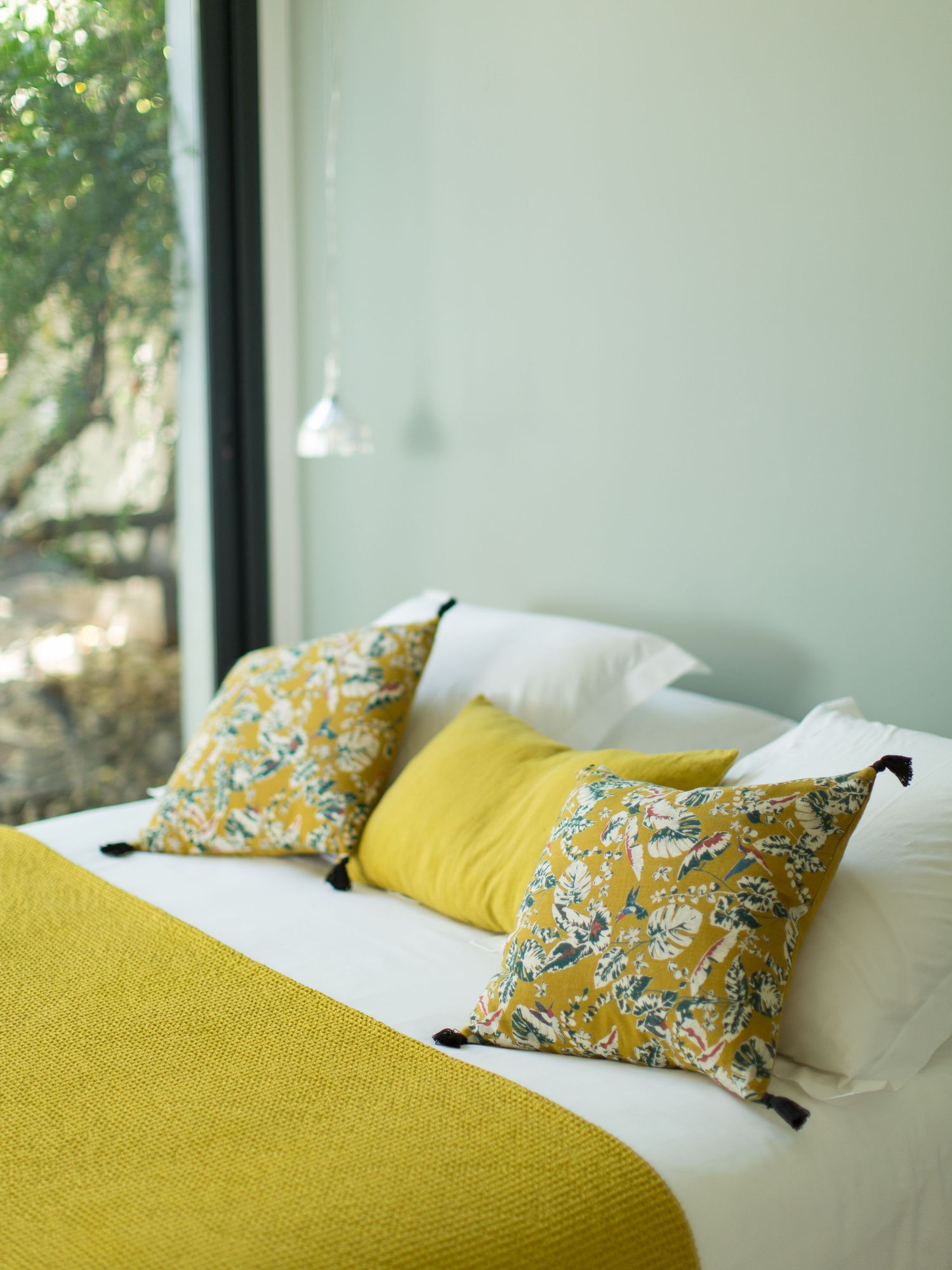 Lit blanc avec jeté de lit en tricot jaune moutarde, coussins à fleurs et jaune uni avec pompons près de la fenêtre donnant sur le jardin.