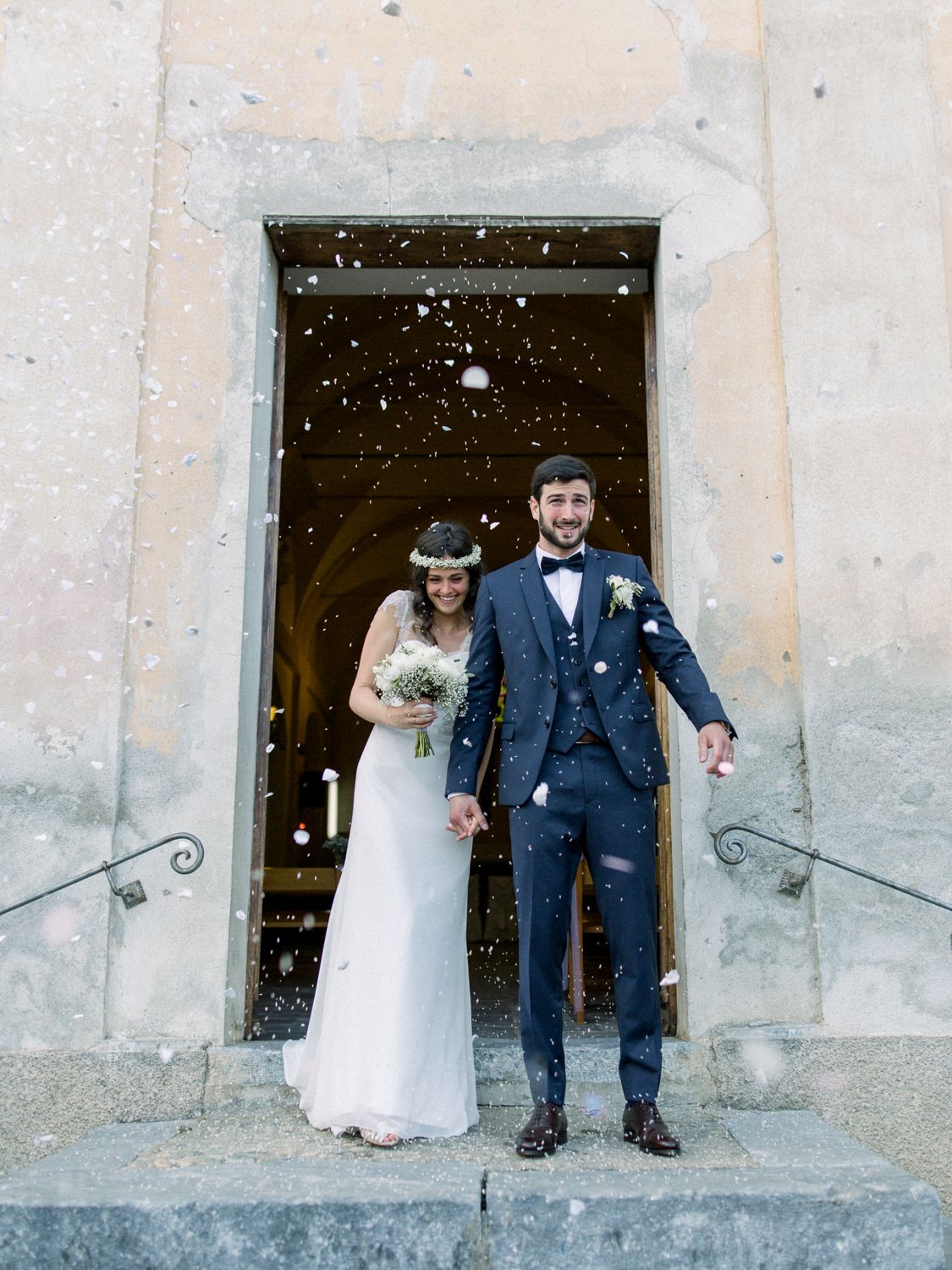 Newlywed couple celebrating outside church entrance with confetti shower, bride in white gown holding bouquet, groom in navy suit.