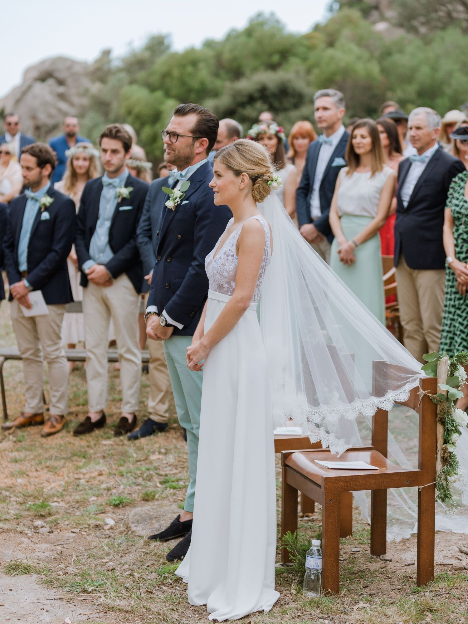 La mariée en robe blanche et voile se tient aux côtés du marié lors de la cérémonie de mariage en plein air, entourée des invités.