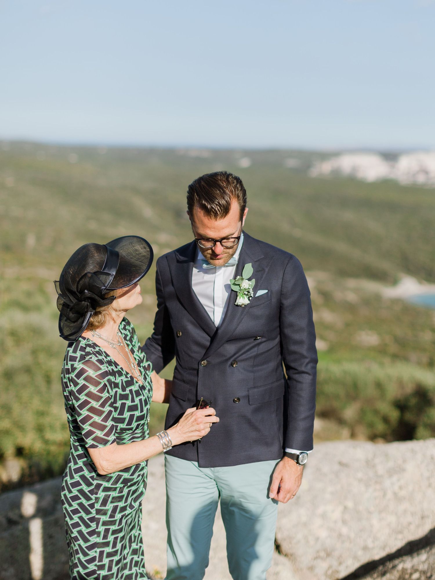 Le marié, vêtu d'un blazer bleu marine et d'un pantalon menthe avec une boutonnière, se tient aux côtés d'une femme élégamment vêtue
