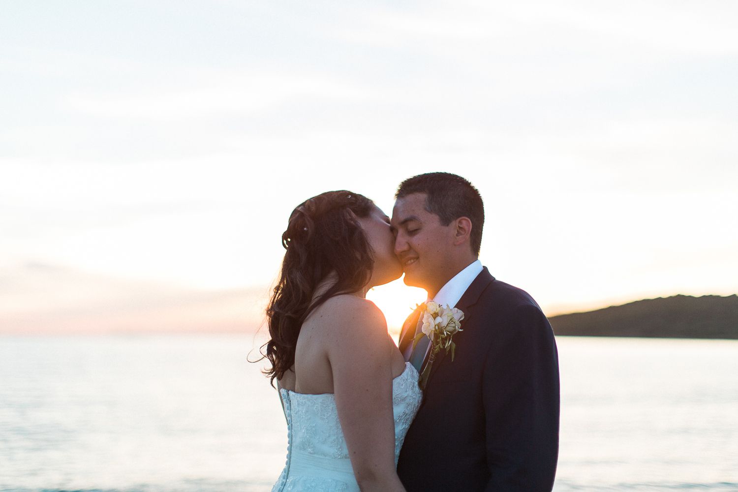 Les mariés échangent un baiser romantique au coucher du soleil, face à l'océan, le jour de leur mariage.