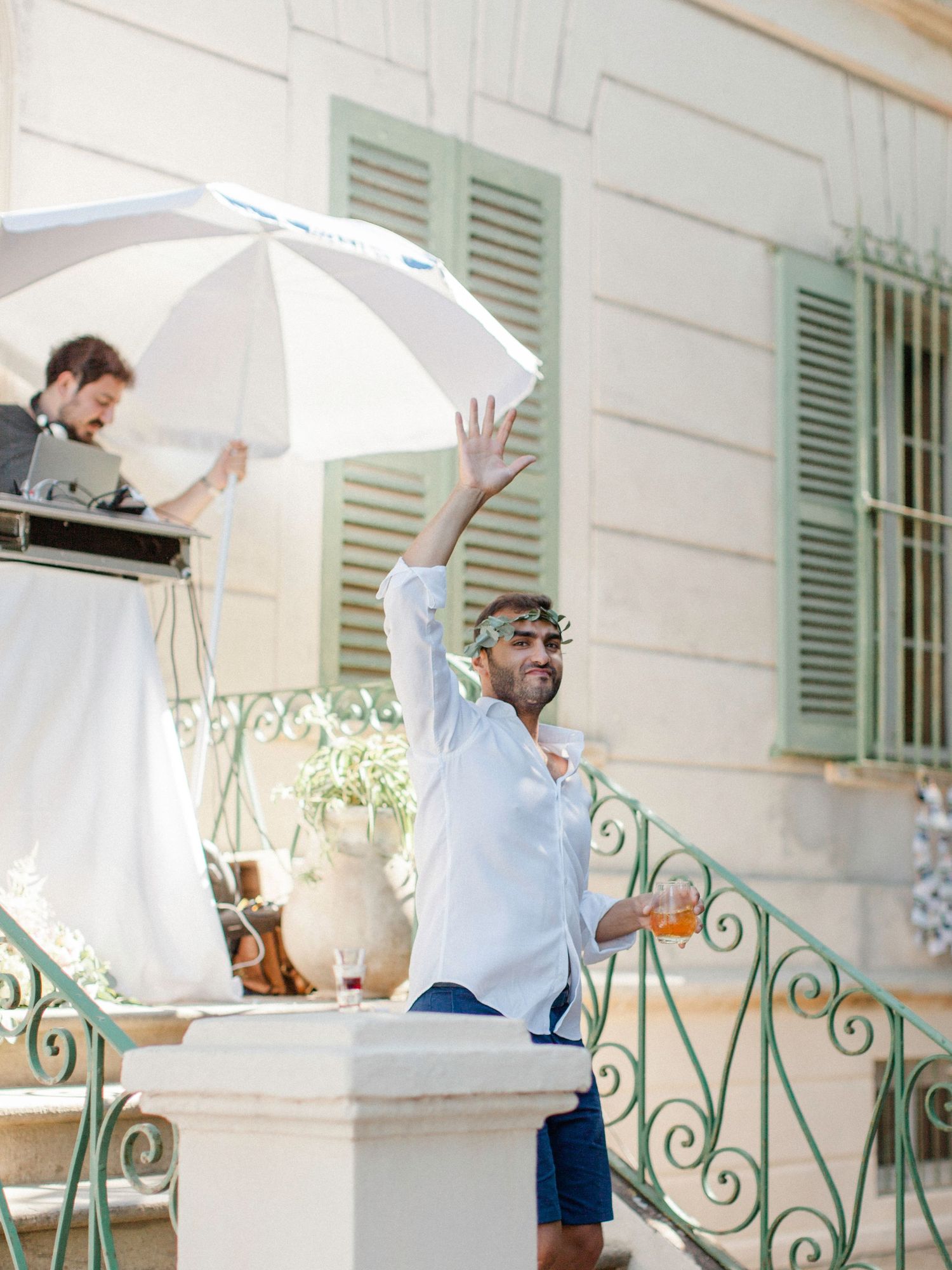 Man in sunglasses raising arm joyfully at outdoor wedding reception near ornate railing and white umbrella.