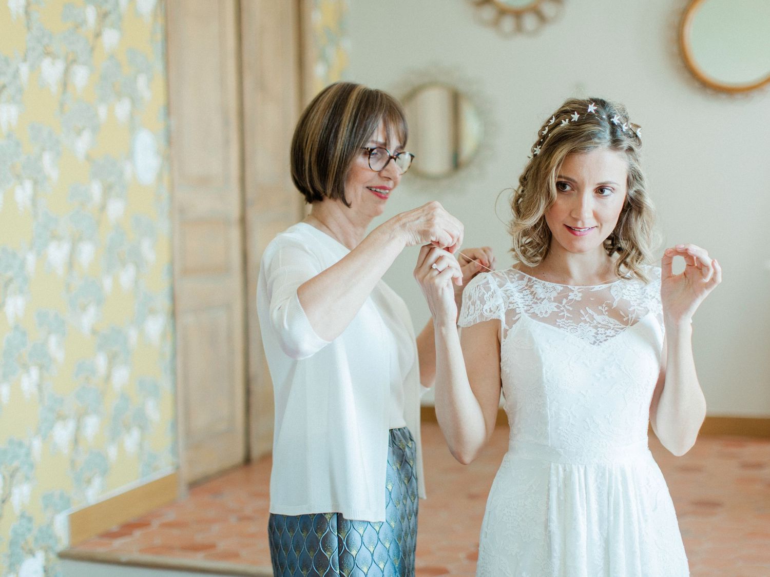A bride in a white lace dress gets help with her necklace from an older woman in a bright, elegantly decorated room.